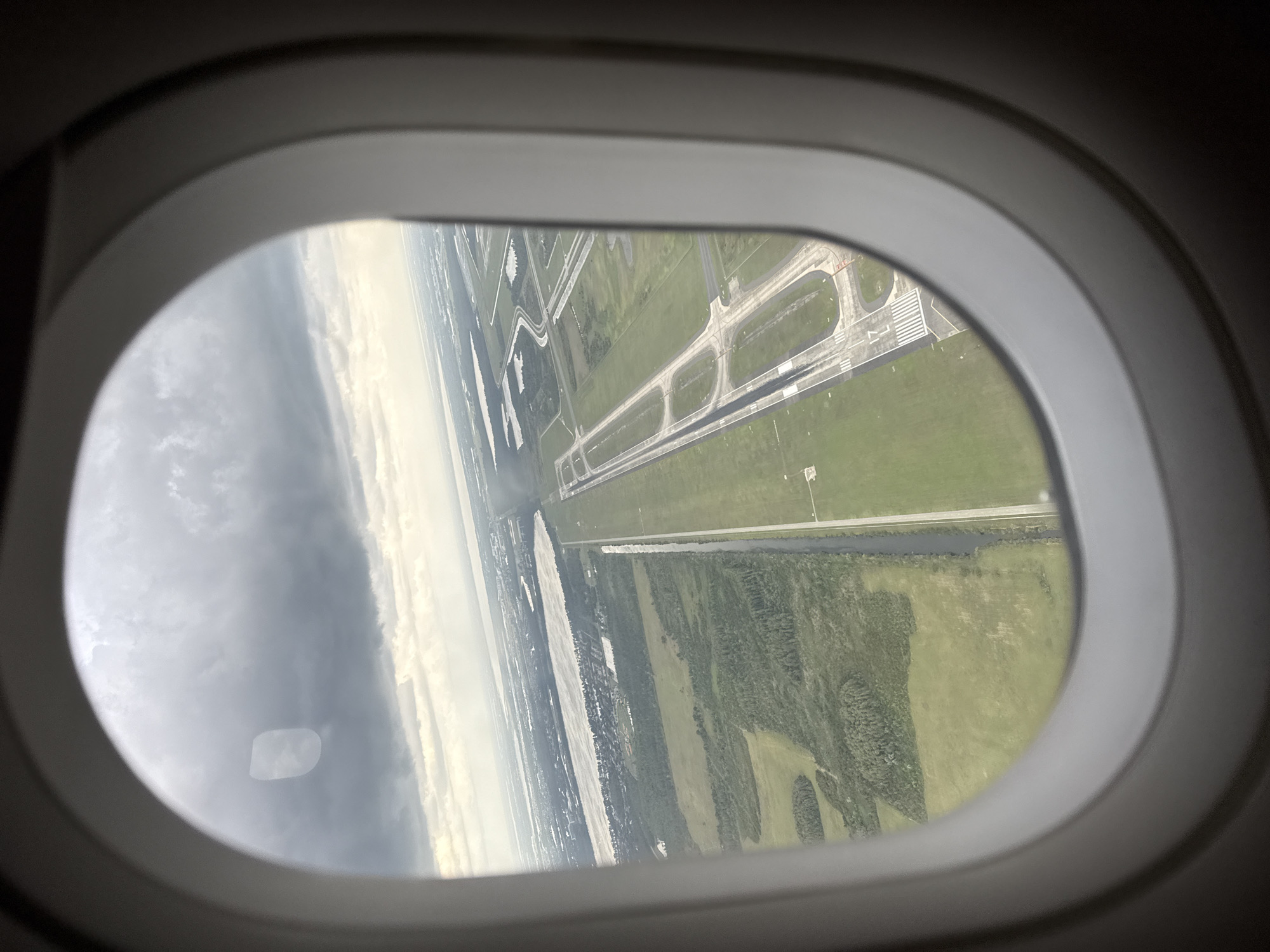 The image captures an aerial view from an airplane window, looking down at an airport runway and surrounding landscape. The dark, curved frame of the airplane window is prominent in the foreground, framing the scene outside.
The view outside is significantly tilted, suggesting the aircraft is banking during takeoff, landing, or a low-altitude maneuver. On the right side of the frame, a large section of an airport runway system is visible. The main runway is clearly marked with white lines, including threshold stripes and the number "17." Several paved taxiways curve and connect to the main runway, all surrounded by extensive areas of vibrant green grass.
Further into the landscape, a distinct body of water, possibly a canal or river, runs parallel to portions of the airport infrastructure. Beyond this, the terrain features patches of dense green trees and more open fields. The sky on the left side of the window appears overcast, with varying shades of gray and white clouds. The lighting indicates it is daytime, possibly under partly cloudy conditions. Given the provided context, this scene depicts an airport in Orlando, United States of America.