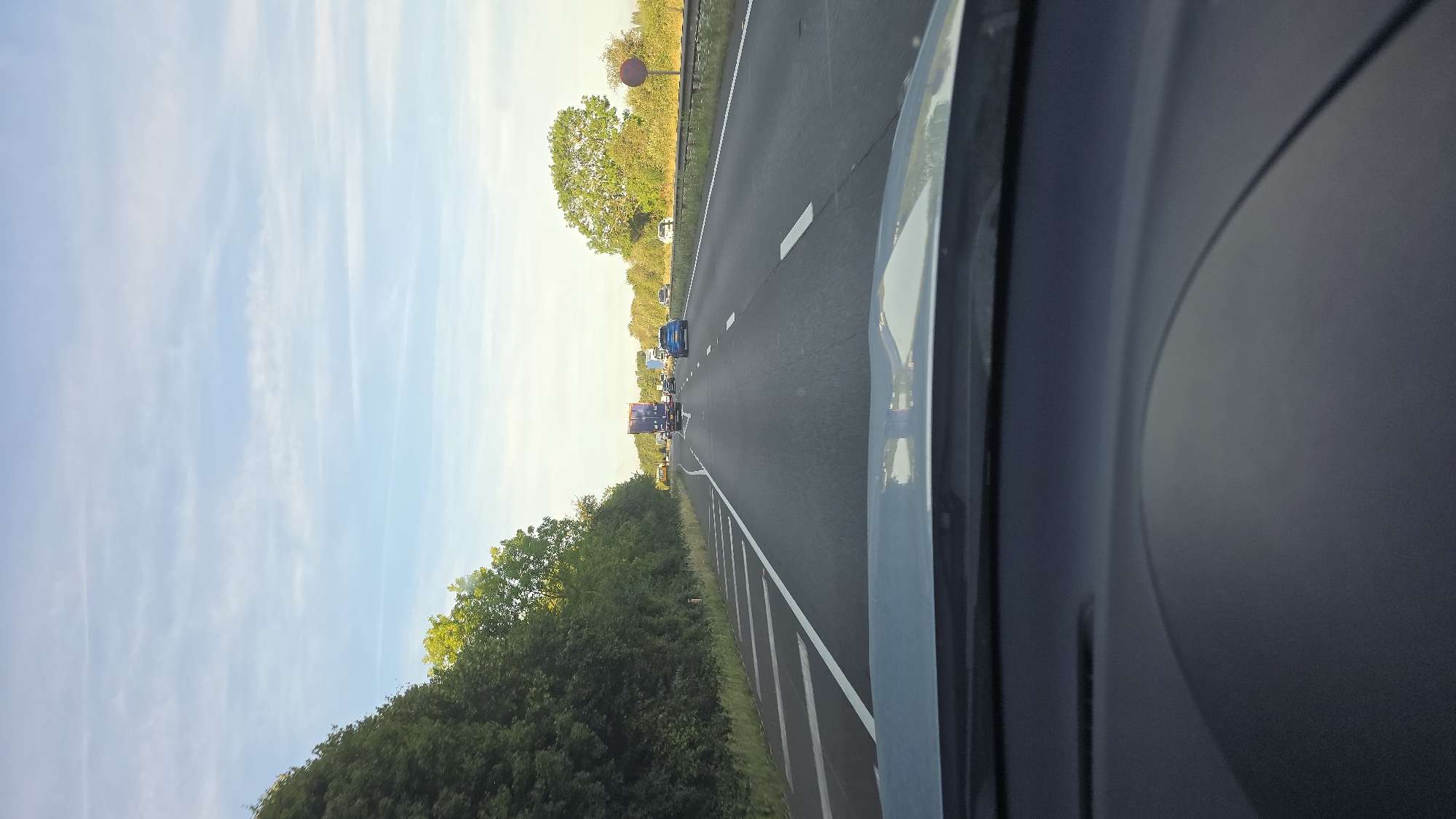 The image depicts a view from the driver's perspective of a car on a highway. The highway has multiple lanes, indicated by white lines. On the left side of the highway, there is a dense line of trees. On the right side, there are trees and a guardrail with a red circular sign. Several vehicles are visible on the highway, including a blue car in the middle lane, a blue truck in the far lane, and other cars and trucks further ahead. The sky is blue with scattered white clouds. The foreground shows the dashboard of the car. The scene suggests a typical daytime drive on a UK highway, potentially within Cherwell District, United Kingdom, given the presence of characteristic highway infrastructure and vegetation.