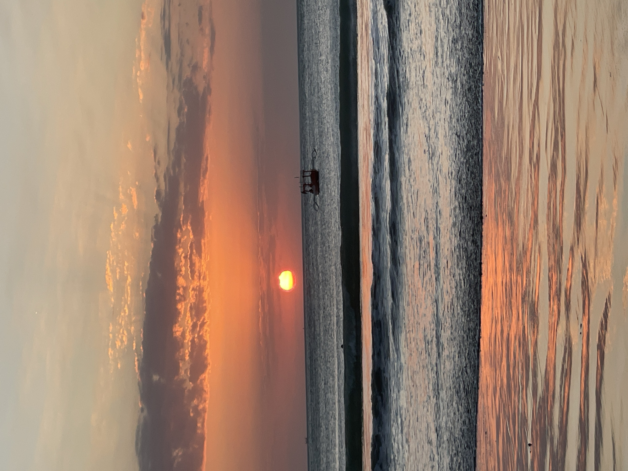 A vibrant sunset paints the sky over the ocean in Kuta Selatan, Indonesia. The sun, a fiery orange orb, is partially submerged below the horizon, casting a warm glow across the water and clouds. A small, red boat with two visible occupants and oars is positioned in the middle of the frame, bobbing gently on the waves. The ocean is calm, with gentle waves rolling towards the shore, reflecting the colors of the sunset. The wet sand in the foreground also mirrors the sky, creating a beautiful, shimmering effect. The overall scene is serene and picturesque, capturing the essence of a tropical evening.