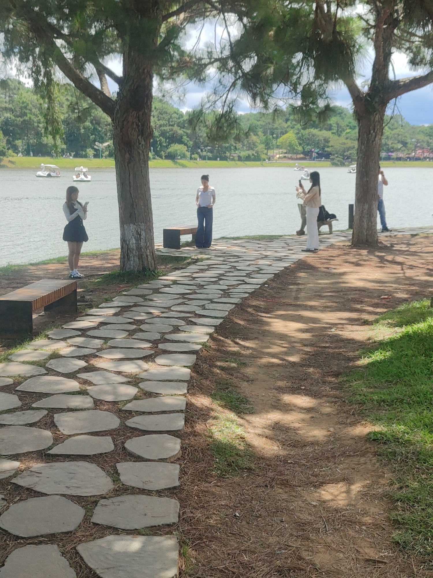 The image depicts a scenic view of Xuan Huong Lake in Da Lat, Vietnam. The foreground features a winding stone pathway bordered by a dirt path and grass, leading towards the lake. Two wooden benches are placed along the pathway. In the middle ground, several individuals are visible near the water's edge. One person, a young woman in a black dress and white top, stands on the left, looking at her phone. Another woman, wearing a white top and blue jeans, stands further down the path, also looking towards the lake. A third person, in a light-colored jacket and white pants, is also near the water, holding a phone. Across the lake, several swan-shaped paddle boats can be seen. The background is filled with lush green trees and hills, under a partly cloudy sky. The overall atmosphere suggests a tranquil day at the popular Da Lat tourist spot.