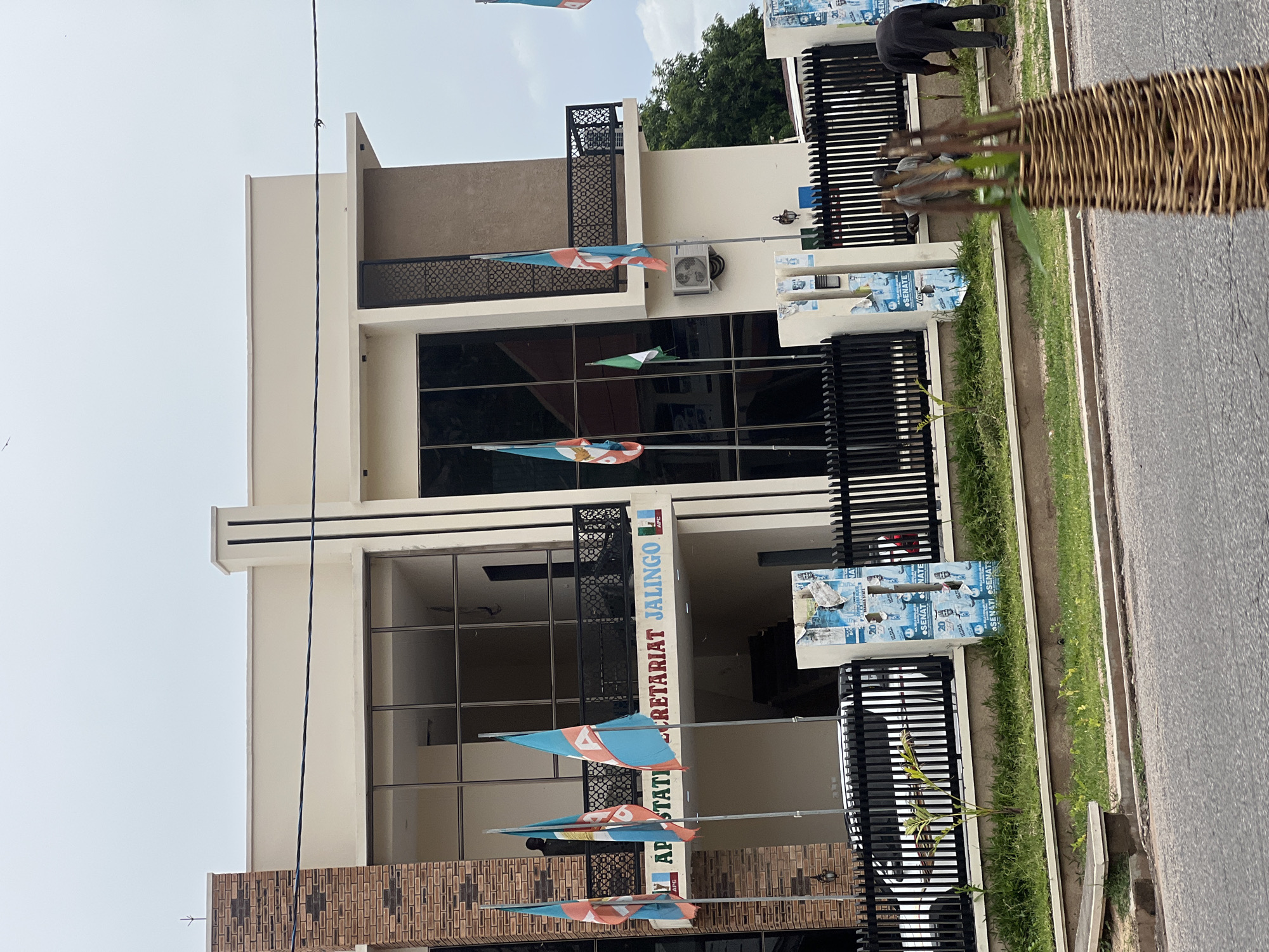 The image depicts the exterior of a modern two-story building, identified by signage as the "APC STATE SECRETARIAT JALINGO." The building features a prominent brick facade on the left and smooth, light-colored plaster on the right. A large, dark-tinted glass facade spans much of the upper level. Several flags are visible, predominantly featuring teal, white, and orange colors, some bearing the acronym "APC." One flag is green and white, possibly the Nigerian flag.
In front of the building, a black metal fence encloses a manicured lawn with some small plants. Behind the fence, a white car is partially visible. Several posters are attached to a support structure near the fence, displaying text such as "SENATE" and "20."
Two individuals are present outside the building. One, wearing a light-colored shirt and dark pants, stands near the entrance. Another person, dressed in dark clothing, is bent over in the grassy area behind the fence, seemingly engaged in gardening or maintenance. The sky is overcast, suggesting a cloudy day. The foreground shows a street with asphalt paving. The scene is located in Jalingo, Nigeria.