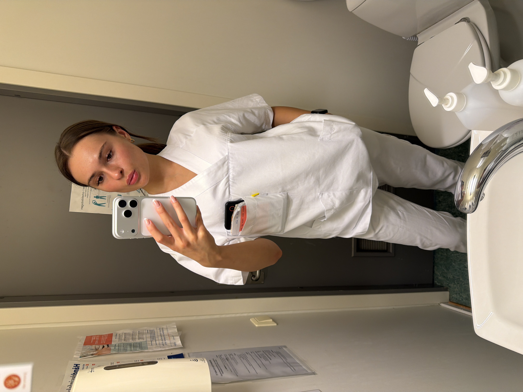 A young woman with brown hair pulled back in a ponytail, wearing white medical scrubs and a watch on her left wrist, is taking a mirror selfie. She has a phone in her right hand, with her left hand resting on her hip. Her scrubs have a pen in the chest pocket and a phone in the side pocket. The background shows a bathroom with a toilet, sink, and a grey door. There is also a dispenser on the wall to the left. The location of Harstad, Norway, is not visible in the image.