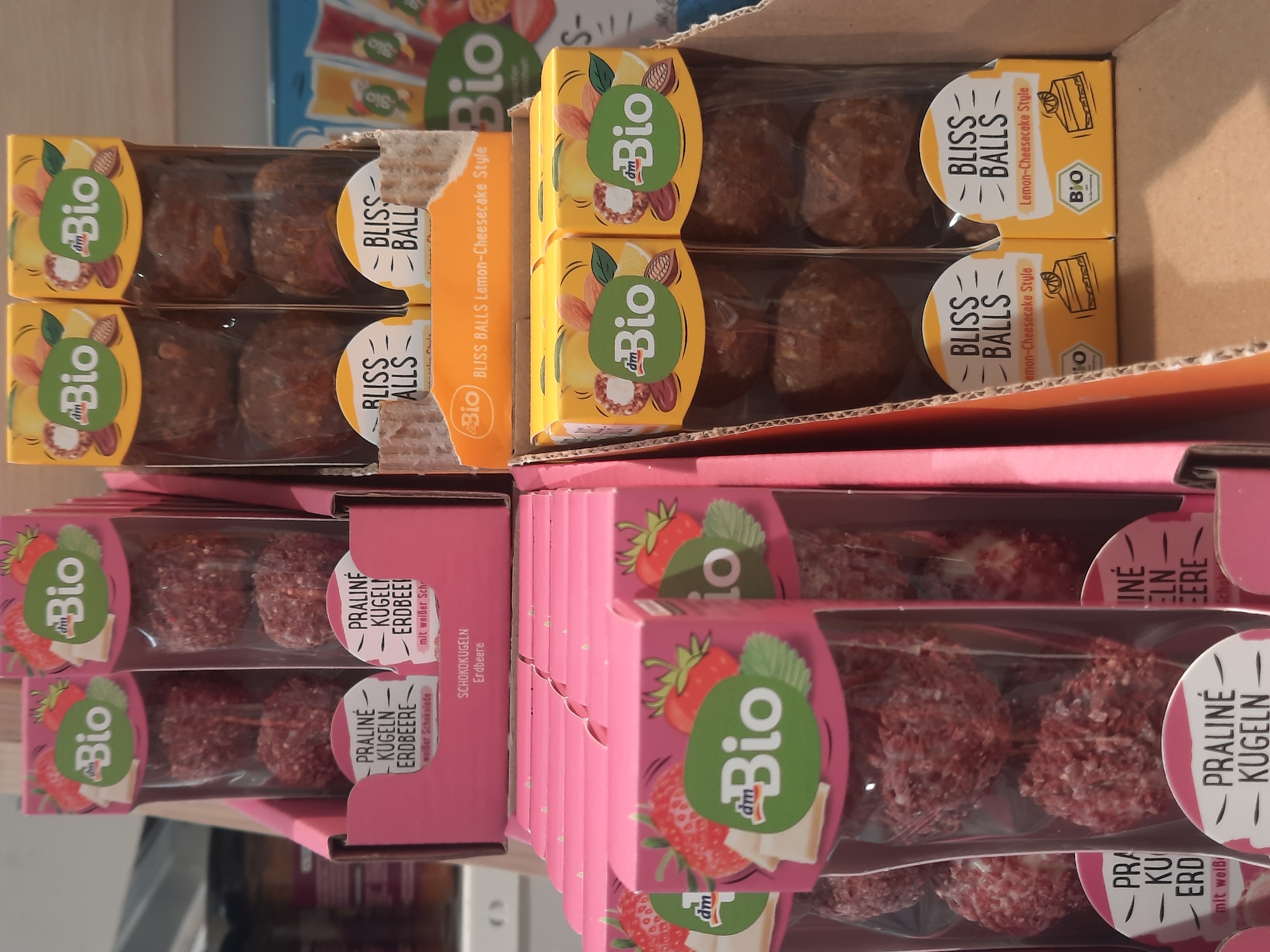 The image shows a retail display of various "dm Bio" branded confectionery products, likely in a supermarket or drugstore, in Rosenheim, Germany. In the foreground and to the left, rows of pink boxes contain what appear to be strawberry-flavored praline balls, each package featuring an illustration of strawberries and white chocolate. To the right, yellow boxes contain "Bliss Balls" in a "Lemon-Cheesecake Style," with illustrations of almonds, lemons, and cheesecake slices. The products are arranged in cardboard trays. The background shows shelves with more products, including what appear to be juice pouches. The lighting is bright, typical of an indoor retail environment. There are no people visible in the image. The overall scene is one of product merchandising.