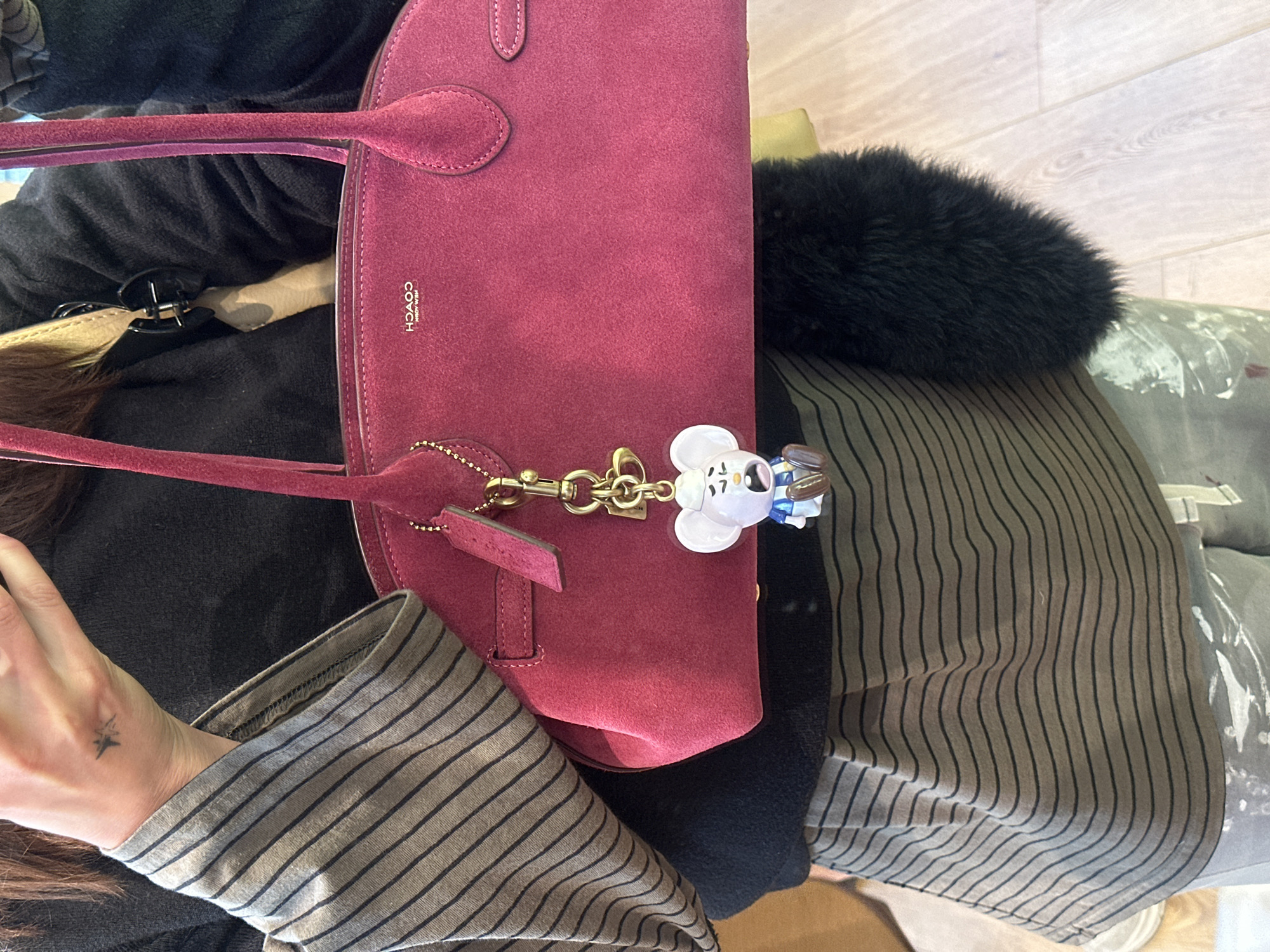 A person is seen seated with a burgundy Coach handbag. The handbag has a decorative charm attached to it. The person is wearing a striped shirt and a black jacket. The location is not discernible and there is no indication that the image was taken in London.