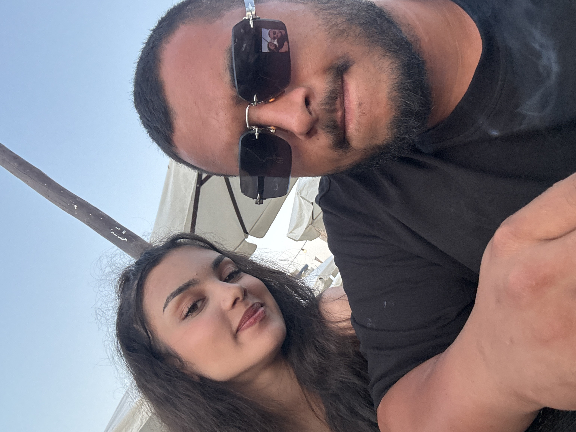 A selfie is taken by a man and a woman in an outdoor setting. The man, wearing sunglasses and a black t-shirt, is in the foreground, with the woman visible in the background to the left. They are positioned against a backdrop of clear blue sky and umbrellas, suggesting a location at an outdoor cafe or seating area. The reflection in the man's sunglasses shows a smaller image of the couple. The image does not provide information to determine the city of The Hague, Netherlands.