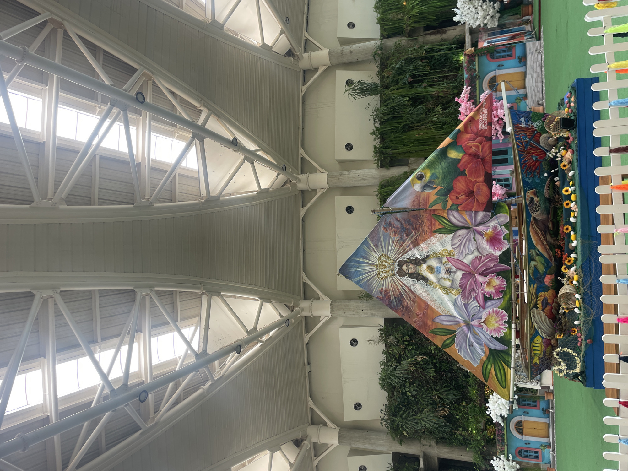 The image depicts a vibrant, decorated boat-shaped float or display, likely part of a festival or public art installation, situated in what appears to be an indoor or covered public space in Pampatar, Venezuela. The float itself is elaborately painted with religious and nature-themed imagery, including a depiction of the Virgin Mary crowned and holding a baby, surrounded by orchids and tropical flowers. A parrot and a sea turtle are also visible on the float's exterior. The boat is adorned with various decorative elements, such as sunflowers, shells, and fishing nets.
Behind the float, a lush green artificial turf covers the ground. A backdrop features colorful, miniature house facades with yellow doors and blue window frames, interspersed with pink flowering trees and white floral decorations. Further back, dense green foliage creates a naturalistic background, suggesting an attempt to simulate an outdoor environment within the covered space.
The overhead structure is a prominent feature, consisting of a large, white, arched roof supported by substantial white beams and columns. The roof incorporates several large skylights, allowing natural light to illuminate the scene. The overall impression is of a festive and artistically curated display designed to be viewed in a public gathering area. There are no people visible in the image.