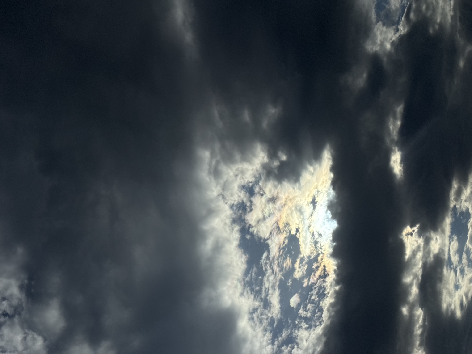 The image shows a sky filled with dark, ominous clouds. Patches of lighter, illuminated clouds are visible through gaps in the darker formations, suggesting the presence of sunlight filtering through. The location in the city of Unknown, Unknown is unknown, with no buildings or other landmarks to help identify the place. The primary subjects are the clouds and the interplay of light and shadow. No people, objects, actions, or interactions are visible in the frame.
