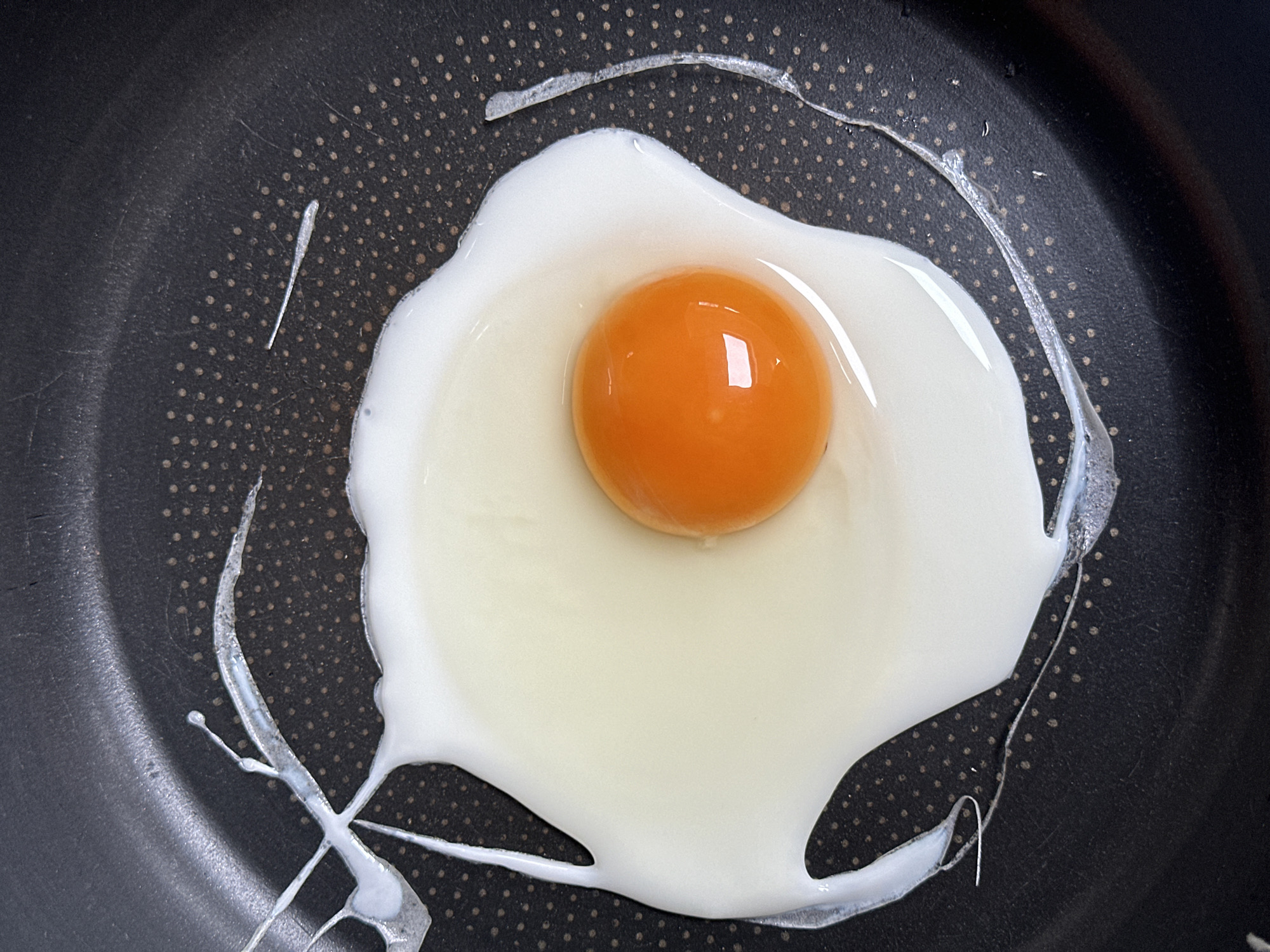 A fried egg is centered in a black frying pan with a speckled surface. The egg consists of a bright orange yolk surrounded by a white albumen. The egg white has spread out and partially coagulated in an irregular shape. A few wisps of uncooked egg white trail off from the main body of the egg onto the pan's surface. No people are visible. There is no information in the image to suggest that this scene is in Tunbridge Wells, United Kingdom.