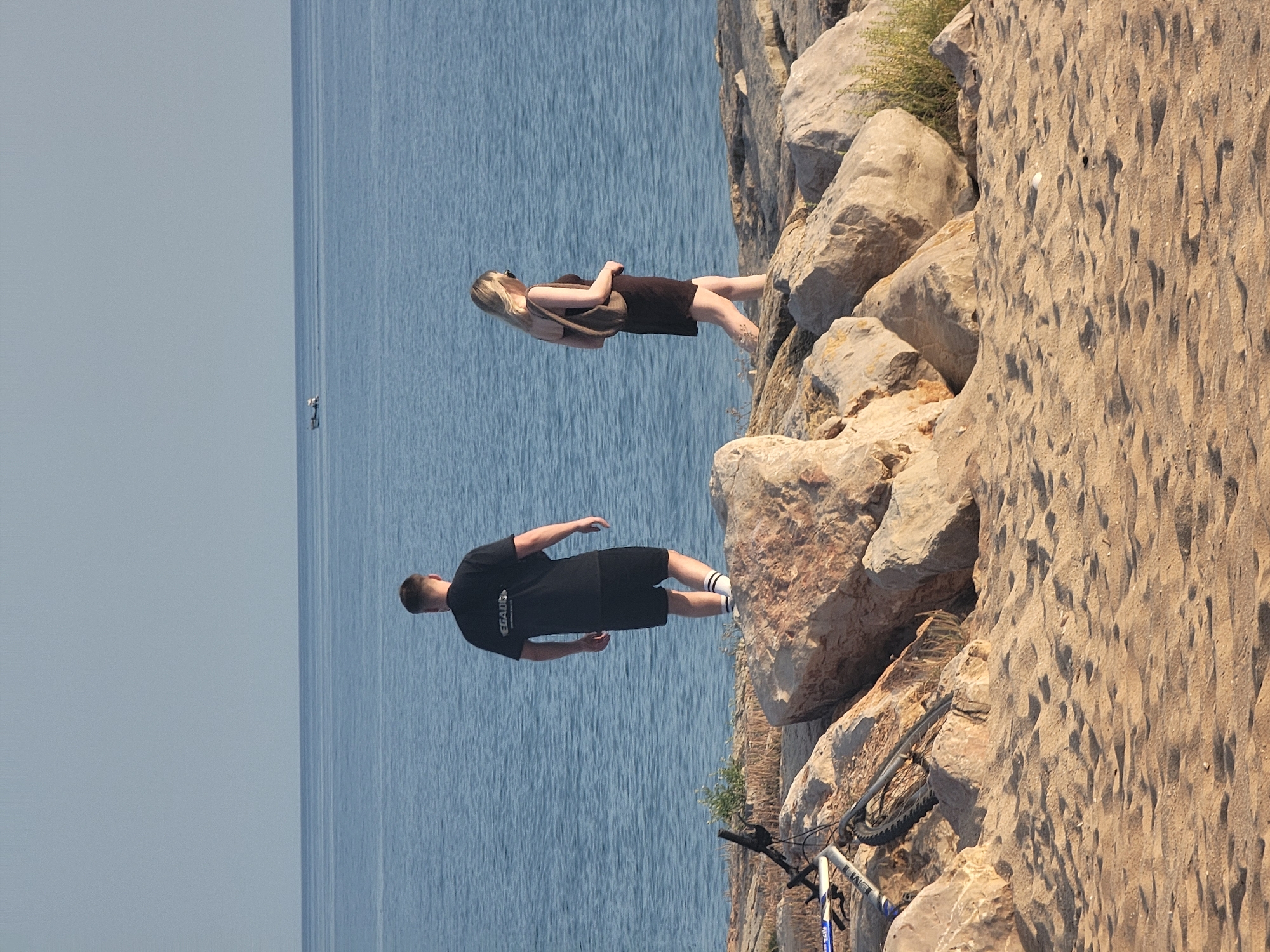 Two people are walking on a rocky shoreline near the ocean in Quarteira, Portugal. On the left, a man wearing a black t-shirt and black shorts with white-striped socks stands on a rock, facing the water. To his right, a woman in a dark-colored dress with a purse slung over her shoulder walks along the rocky area. A bicycle is lying on its side on the sand and rocks in the foreground, to the left of the man. The ocean stretches out to the horizon under a clear, pale sky. A small boat is visible in the distance on the water. The ground is covered in sand and large, light-colored rocks.