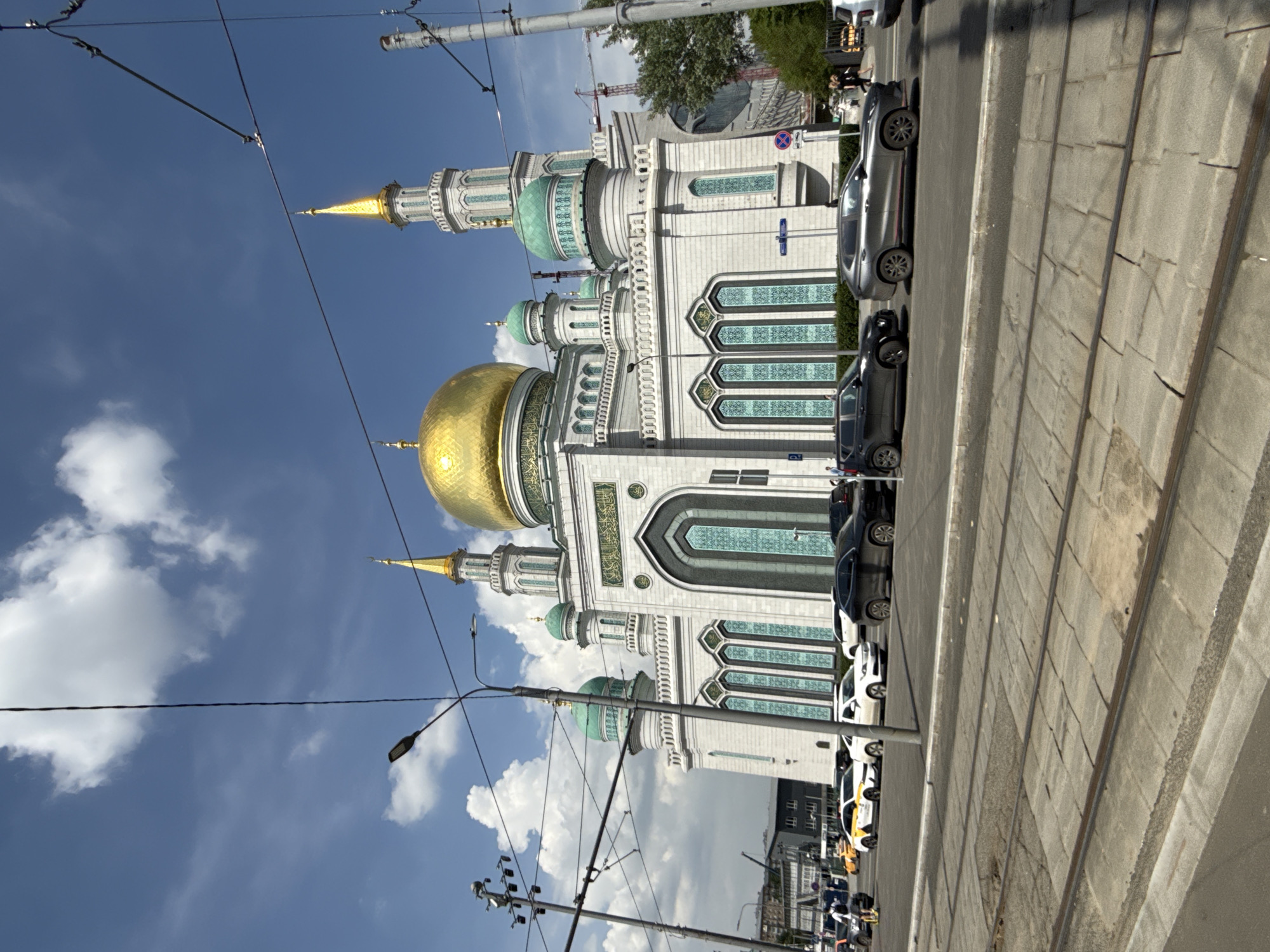 The image depicts the Moscow Cathedral Mosque in Moscow, Russia, from a street-level perspective. The mosque, a large white structure with a prominent golden dome and several turquoise domes and minarets, dominates the view. The building's facade features arched windows with intricate geometric designs. Several cars are parked along the right side of the image along the roadway. Overhead electrical wires and streetlights are visible against the blue sky with scattered clouds. In the lower right corner of the image, a sidewalk and railroad tracks are visible.