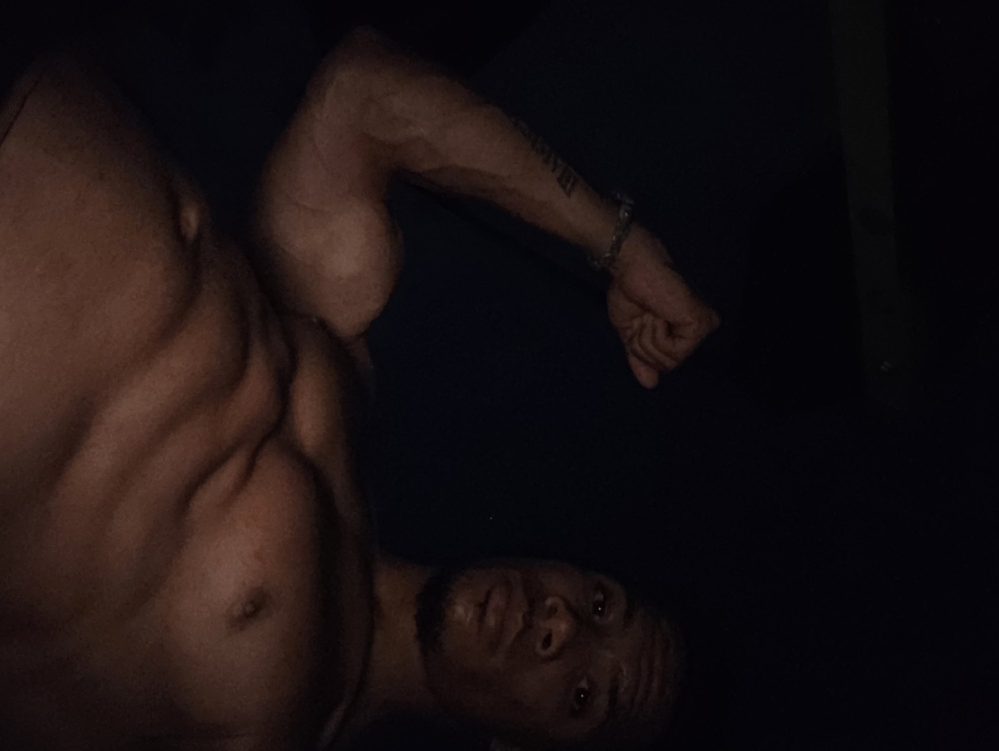 In a dimly lit indoor environment, a muscular individual with dark skin is posing, showcasing their physique. The subject's left arm is flexed, revealing well-defined biceps and musculature. A bracelet is visible on their left wrist, and a tattoo extends along their forearm. The individual's chest and abdomen are also visible, with prominent abdominal muscles. Their face is turned towards the camera, with dark eyes, a straight nose, and a light beard. The background is dark and indistinct, suggesting a low-light setting. The scene does not provide any information that would allow for its location to be identified as Varna, Bulgaria.