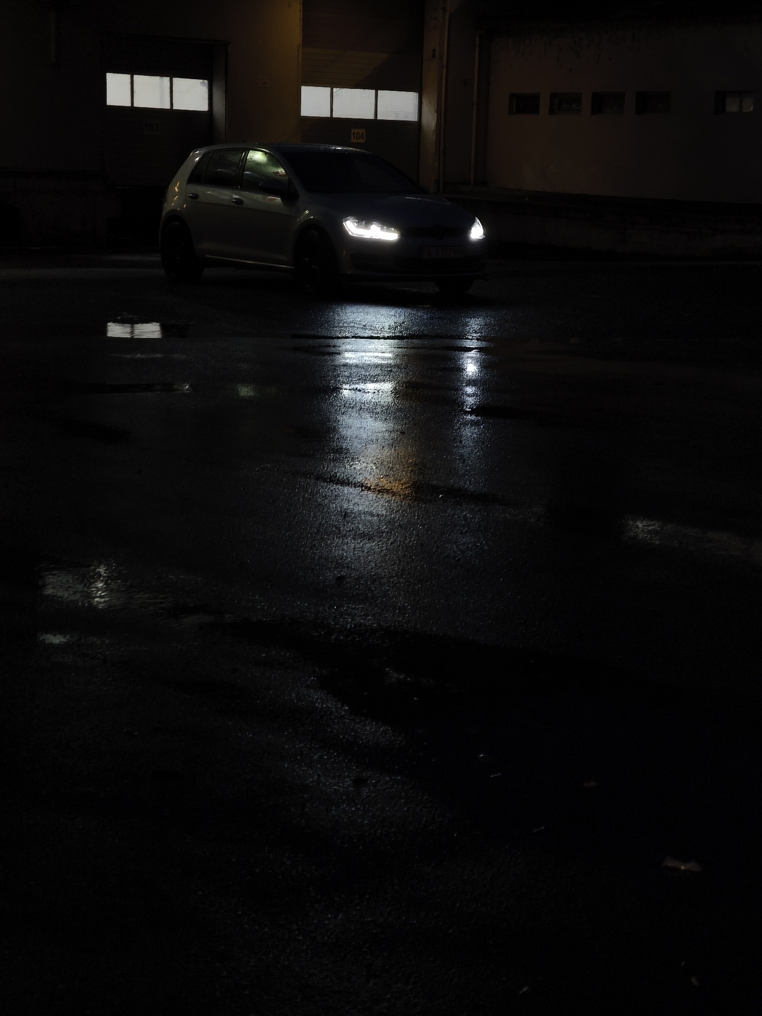 The image captures a dark, nighttime scene in Burgas, Bulgaria, featuring a light-colored Volkswagen Golf with its headlights illuminated. The car, possibly silver or white, is angled slightly towards the viewer, showcasing its bright LED headlamps which cast strong reflections on the wet asphalt surface. The car is equipped with dark-colored wheels, and a partial view of its Bulgarian license plate, "A 5131 HA", is visible on the front bumper.
The ground is a dark, wet asphalt, highly reflective due to moisture, suggesting recent rain. Streaks of light from the car's headlights and reflections of the building's lights create glistening patterns across the damp surface, with small puddles enhancing the reflective quality.
In the background, a dark, industrial-style building is visible. It features several rectangular windows or panels that are lit from within, emitting a soft yellow glow. Two large garage-style doors are also part of the building's facade, with the numbers "103" and "104" clearly marked above them. The overall scene is characterized by deep shadows and stark contrasts, conveying a moody and atmospheric impression of an urban or industrial area after dark.