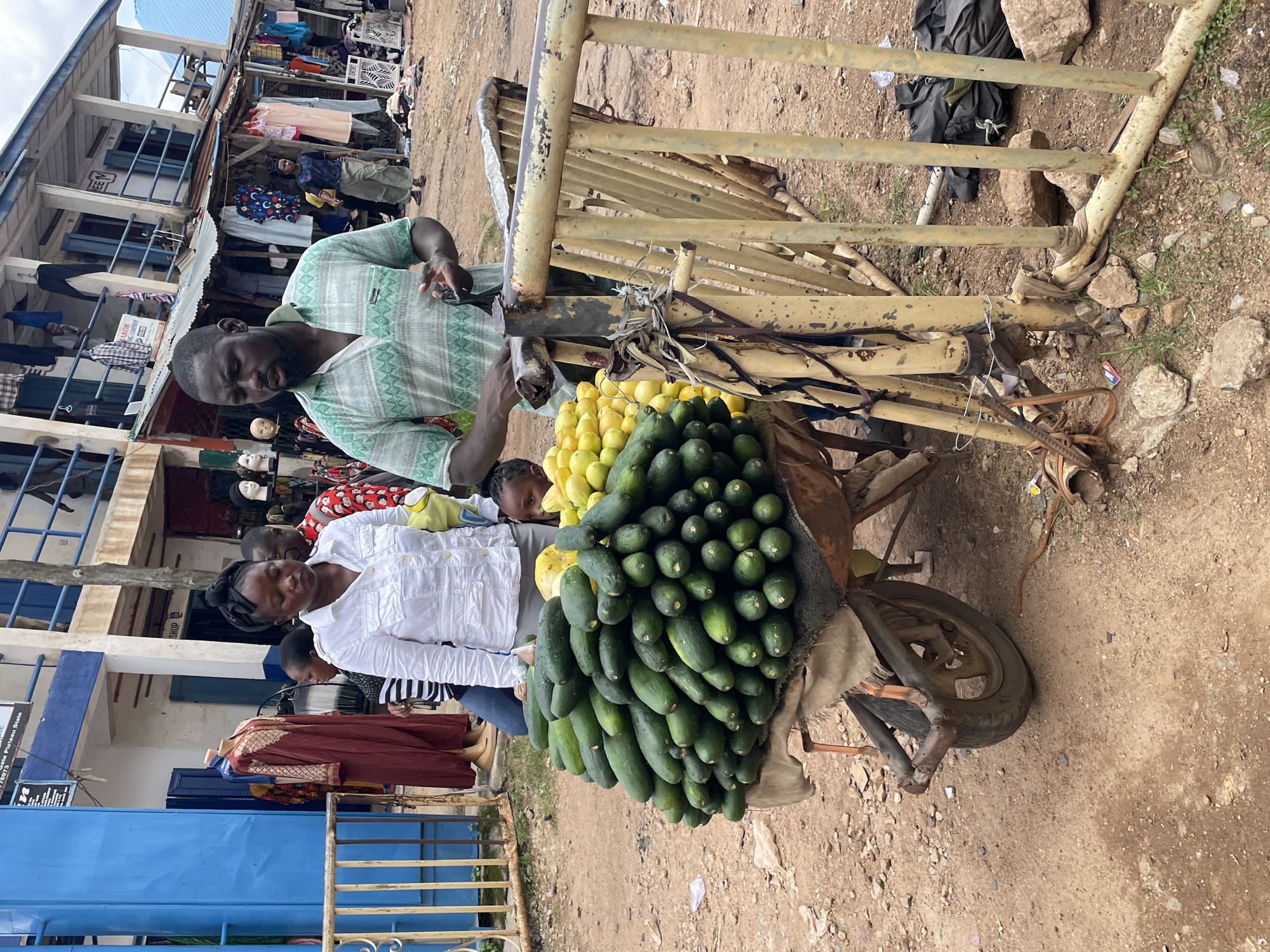This image depicts a street market scene in Jos, Nigeria. A man wearing a green and white patterned shirt is standing next to a wheelbarrow overflowing with cucumbers. Beside the cucumbers, there is a pile of yellow fruits, possibly lemons or limes. A woman in a white shirt stands behind the wheelbarrow, and a young child is visible looking out from behind her. In the background, there are market stalls with clothing and various goods displayed, and other people are present, some browsing and some working. The ground is dirt and gravel, and a yellow metal barrier runs along the right side of the frame. The overall impression is of a busy, everyday market environment.