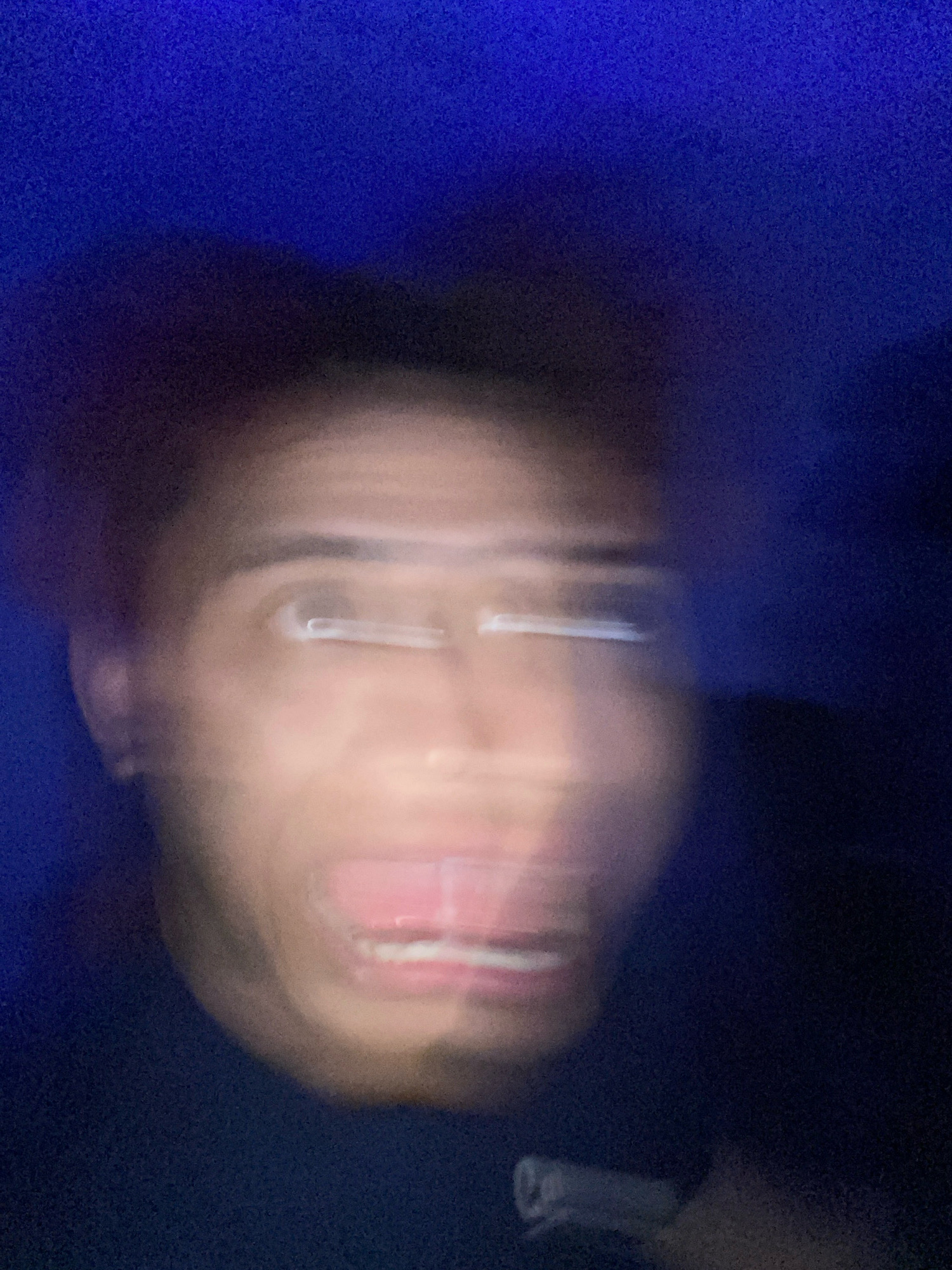 The image is a blurry, close-up photograph of a person's face. The person appears to be looking directly at the camera with their mouth open, revealing teeth. Their eyes are also visible, but are blurred. The background is a dark, out-of-focus blue, suggesting an indoor setting with artificial lighting. There are no discernible landmarks or elements that would indicate a specific location within Klurak, Indonesia. The focus is entirely on the facial features, which are distorted by motion blur.