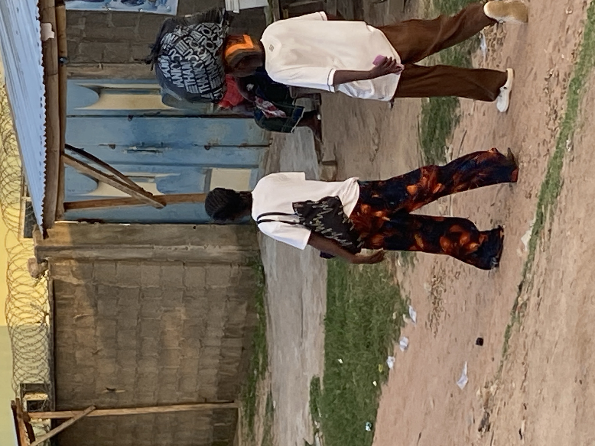 In Jauro Yinu, Nigeria, two individuals are walking along a dirt path. The person on the left, viewed from behind, wears a white t-shirt and vibrantly patterned pants. They carry a dark handbag with a white pattern on their left shoulder. The person on the right, also viewed from behind, is dressed in a white t-shirt and brown trousers. They are carrying a large black bag on their head, which appears to be filled with items and has white text printed on it. In the background, a rough concrete wall topped with barbed wire runs horizontally. Beyond the wall, a corrugated metal roof is visible. A blue metal door stands in a wooden frame, with a person seated in front of it, partially obscured. The ground is a mixture of dirt and sparse patches of green grass, with scattered debris. The lighting suggests it may be late afternoon or early morning.