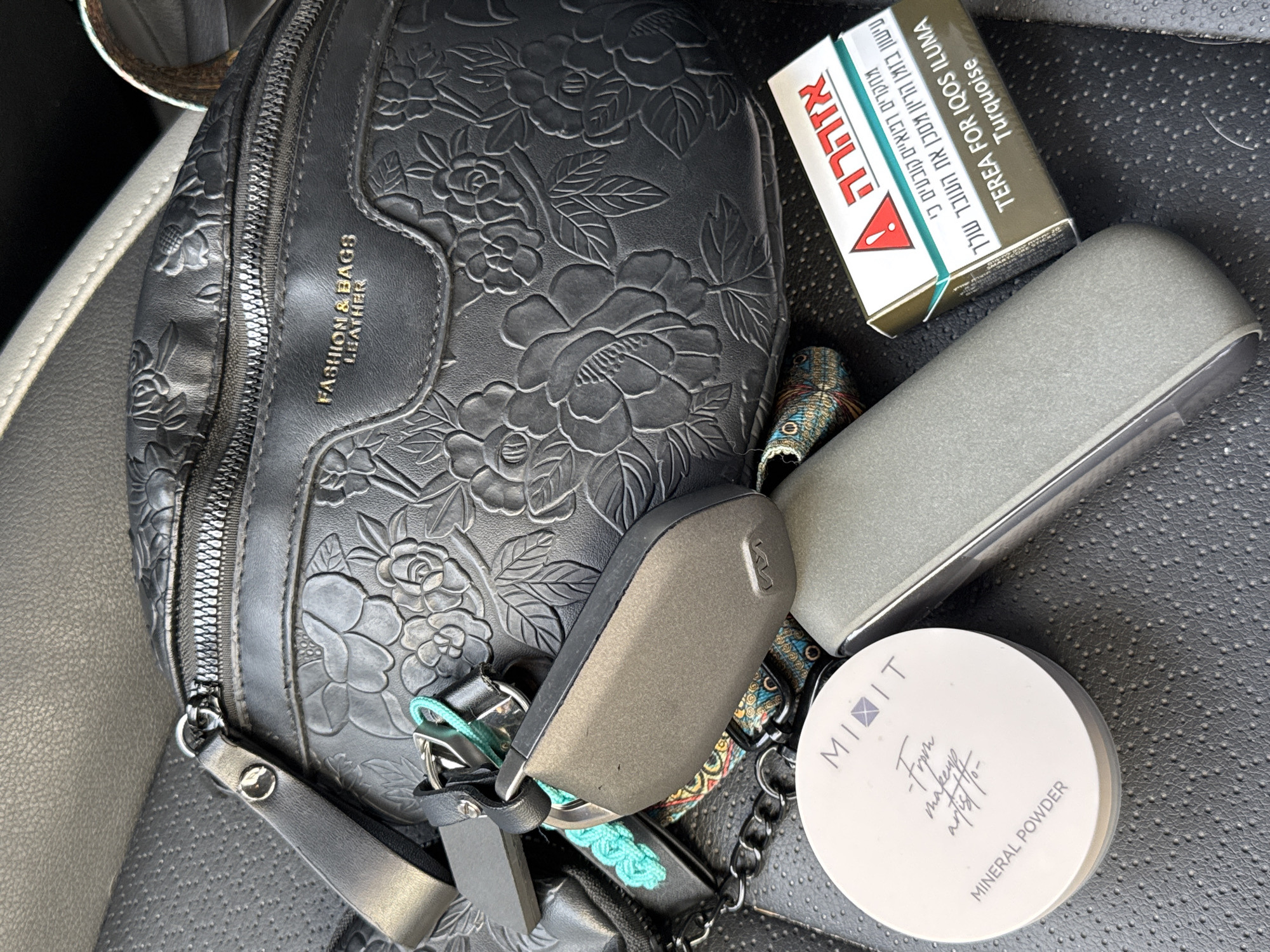 The image shows a black leather purse with a floral pattern sitting on a black leather seat. Next to the purse are a pack of TEREA FOR IQOS ILUMA Turquoise cigarettes with Hebrew text, a gray eyeglasses case, a white round container of MIXIT Mineral Powder makeup, and car keys. There is no indication of the image location being Tel-Aviv, Israel, beyond the Hebrew text on the cigarette pack.
