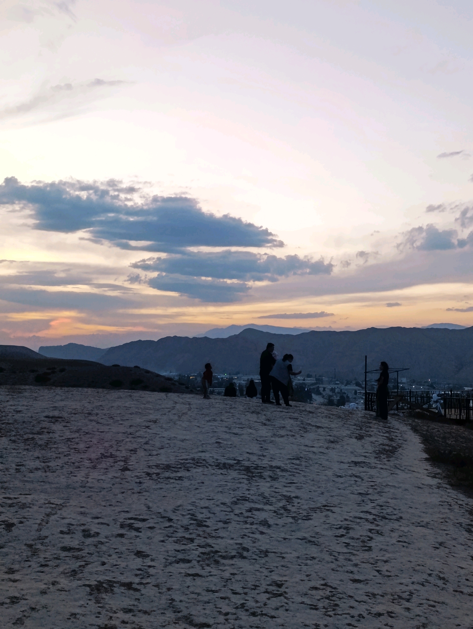 The image captures a panoramic view of Naryn City, Kyrgyzstan, during a sunset. The sky is filled with dramatic clouds, ranging in color from pale yellow and orange near the horizon to shades of grey and blue higher up. In the foreground, a sandy, uneven terrain slopes downwards towards the city. Several silhouetted figures are scattered across this foreground, suggesting they are observing the sunset or the cityscape below.
On the left side of the frame, a child stands facing away from the camera. Further to the right, three individuals are grouped together; one appears to be taking a photograph with a mobile device. To the far right, another silhouetted figure stands looking out over the landscape. In the middle ground, the city of Naryn is visible as a collection of buildings nestled within a valley, with mountains forming a majestic backdrop. The mountains themselves are layered, with the closest ones appearing dark and silhouetted against the softer hues of the sky, while distant peaks fade into a hazy, purplish tone. A fence-like structure is partially visible on the right edge of the image, suggesting a viewing platform or a boundary. The overall atmosphere is serene and contemplative, typical of a sunset vista.