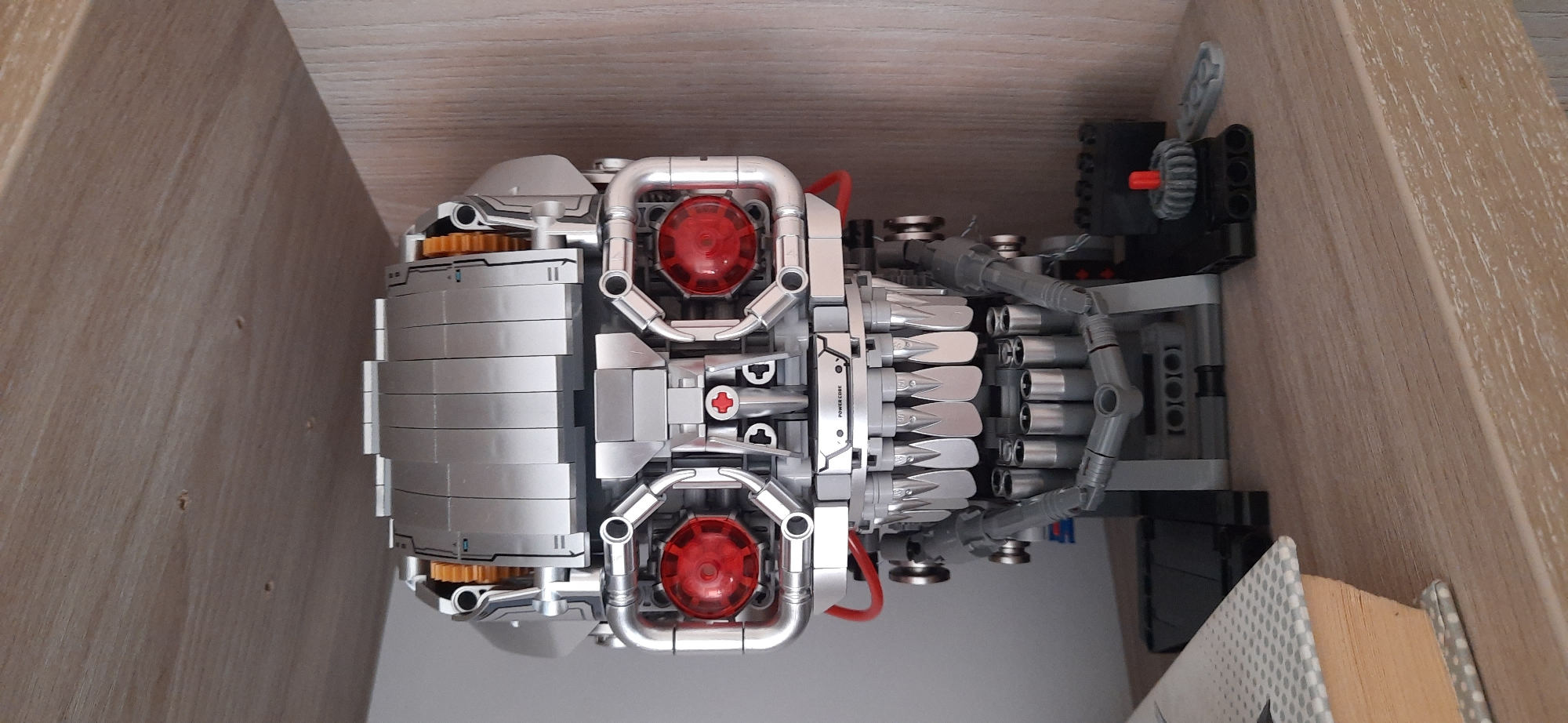 A Lego model of a Terminator skull sits on a wooden shelf. The model is metallic silver and features red circular "eyes" and a row of sharp, metallic teeth. The skull is mounted on a black Lego base with some additional Lego pieces to the right of the base. To the left of the skull, the corner of a book with a grey and white polka-dot cover is visible. The background consists of a wooden shelf and a light-colored wall. The image was taken indoors, in an apartment in Dnipro, Ukraine.
