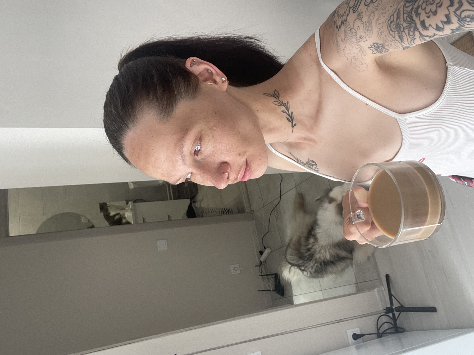 A woman with dark hair pulled back in a ponytail is holding a glass mug filled with a light brown liquid, likely coffee or tea with milk. She has multiple piercings in her ear and nose, and visible tattoos on her neck and arm. She is wearing a white ribbed tank top. A fluffy gray and white dog is visible behind her. The scene appears to be inside a home, with a bathroom visible in the background. The exact location in Starki, Russia cannot be determined from this image alone.