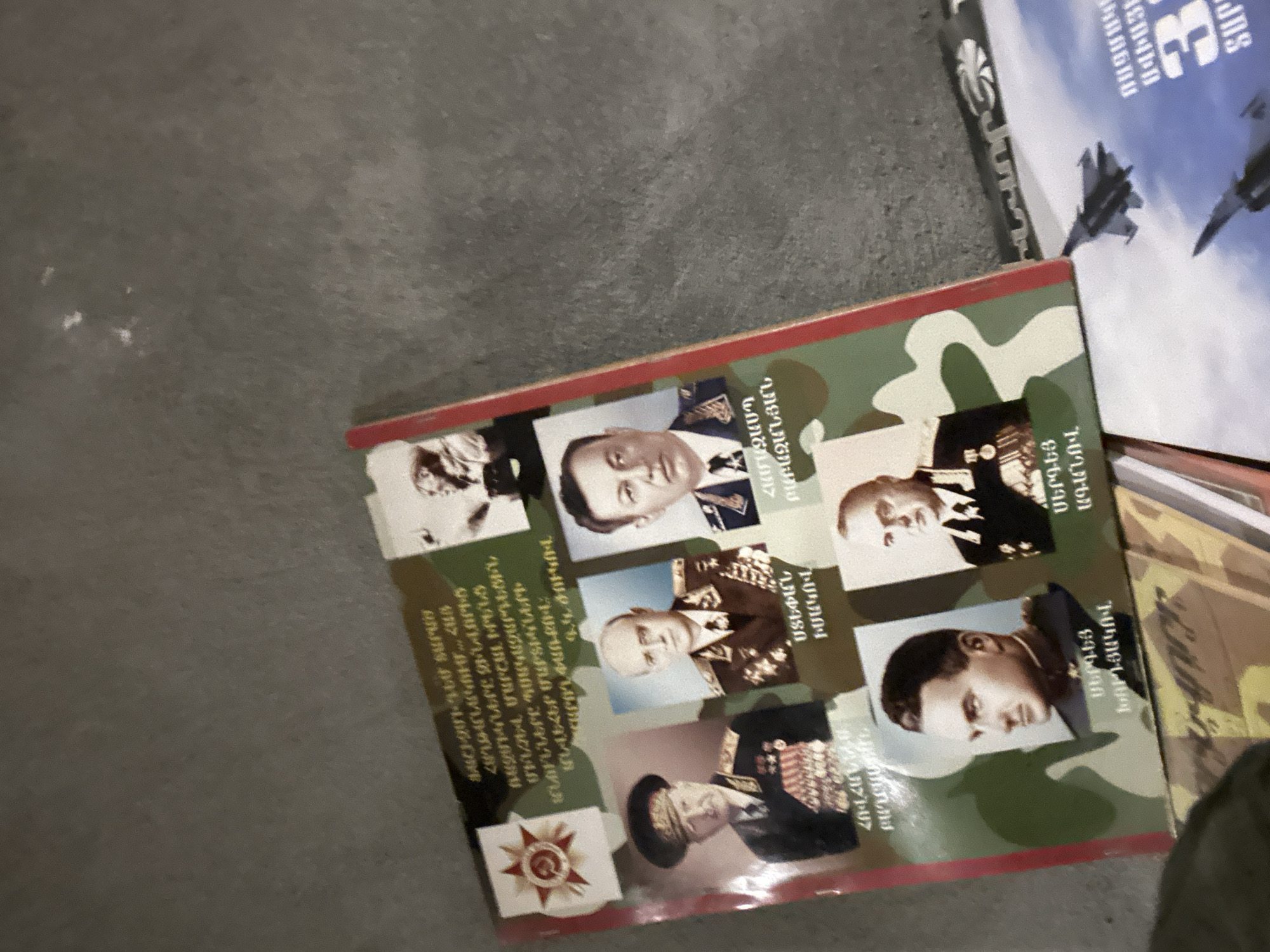 The image shows a book cover featuring portraits of military figures with Armenian text, placed on a surface alongside another book depicting fighter jets. The book cover includes names like Stepan Isakov, Hamazasp Babazanyan, Sergey Khudyakov, and Sergey Aganov. The background features a camouflage pattern and an emblem resembling a star or medal. The context suggests it's likely a book about military history or biographies of military personnel.