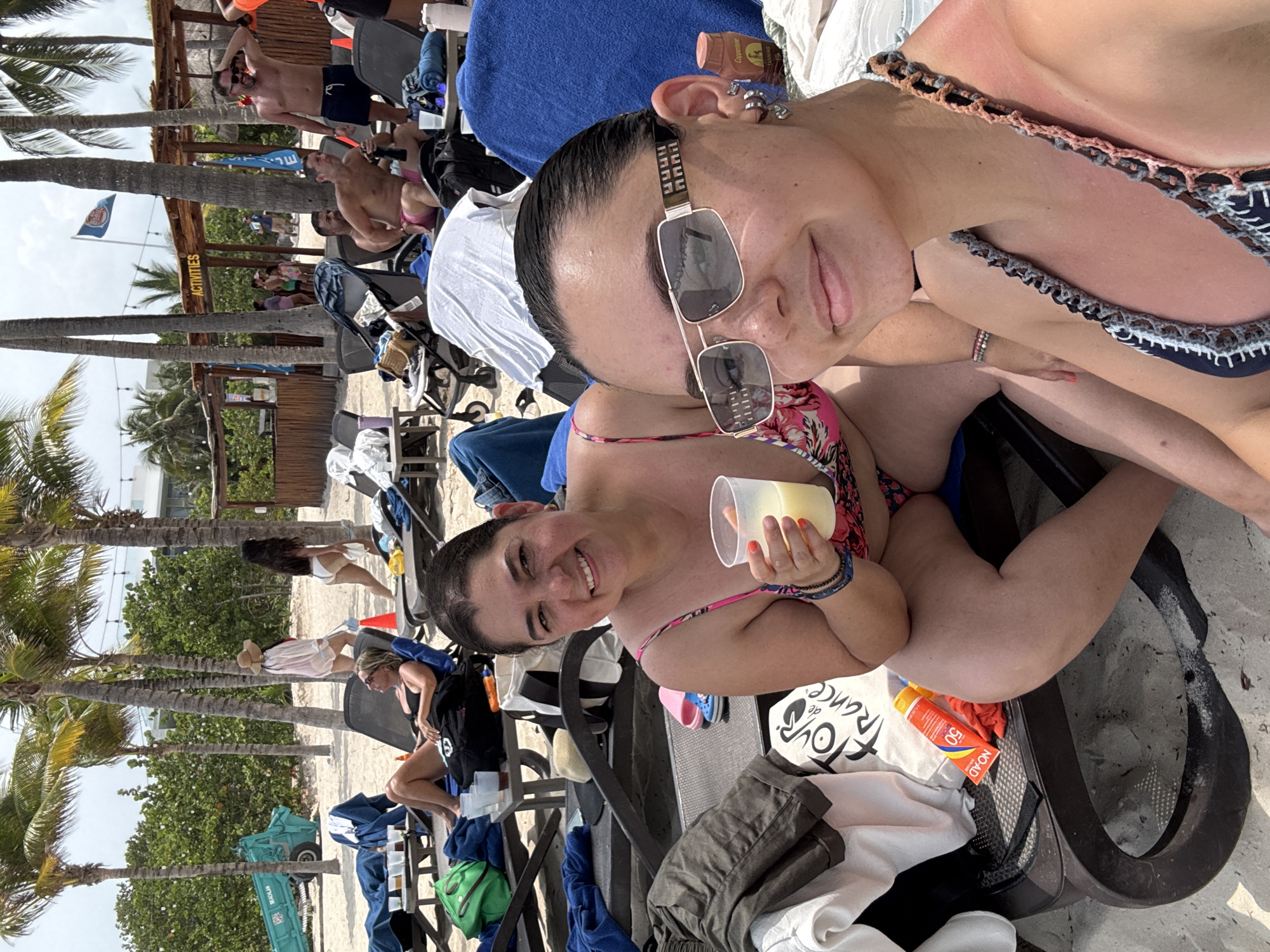 Two women pose in beach chairs at a beach in Playa del Carmen, Mexico. One woman holds a drink. Palm trees are present. Beachgoers are visible in the background. An 'Activities' sign is also visible.