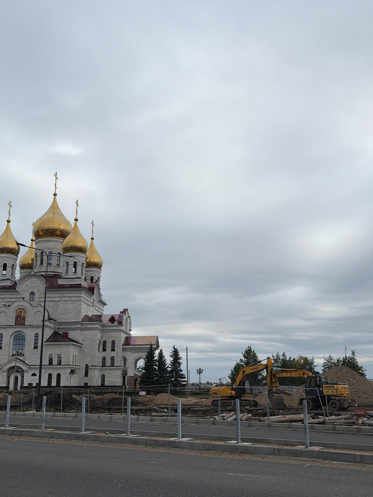 A large, white Orthodox church with multiple golden domes and crosses is visible on the left side of the frame. Construction equipment, including two yellow excavators and a pile of dirt, is present on the right side. A fence separates the construction area from a paved road in the foreground. The sky is overcast and gray. The church appears to be the Holy Trinity Cathedral in Arkhangelsk, Russia, as suggested by its distinctive architecture and the surrounding environment.