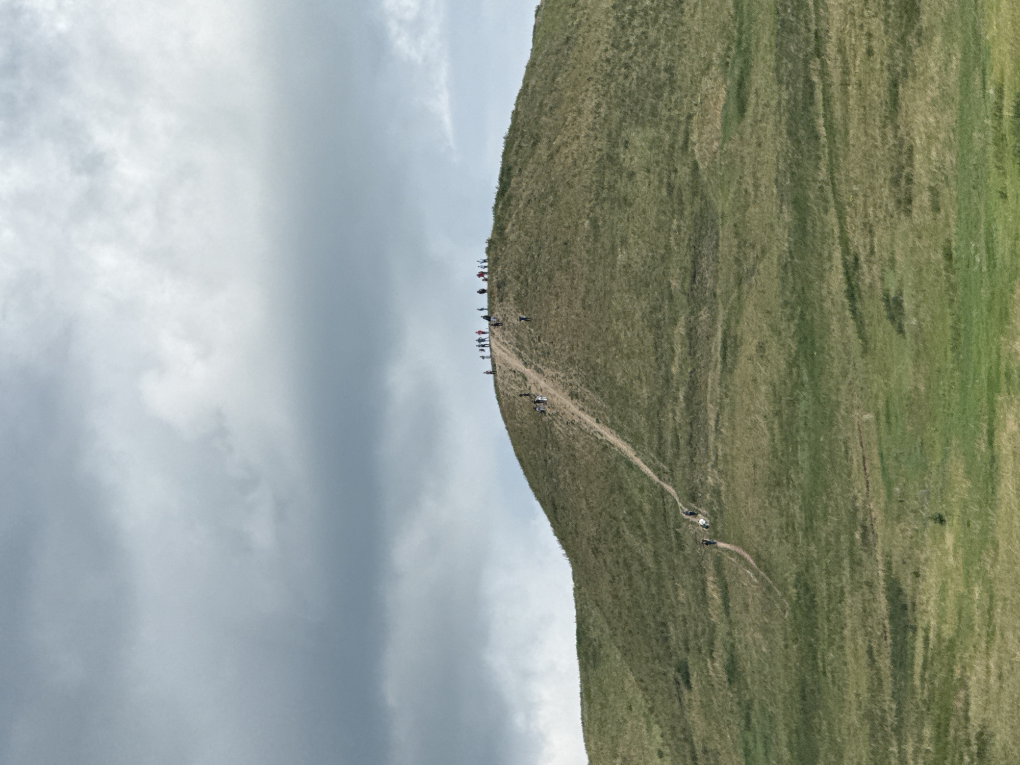 The image depicts a grassy hill with a winding dirt path leading to its summit. A group of people are scattered along the path and at the top, appearing to be hiking or exploring. The sky above is overcast with dramatic, dark clouds, suggesting a moody or potentially stormy atmosphere. The foreground consists of more rolling, green terrain. The overall impression is of an outdoor recreational activity in a natural, somewhat rugged landscape. The location is stated to be in the city of Nuryevo, Russia.
