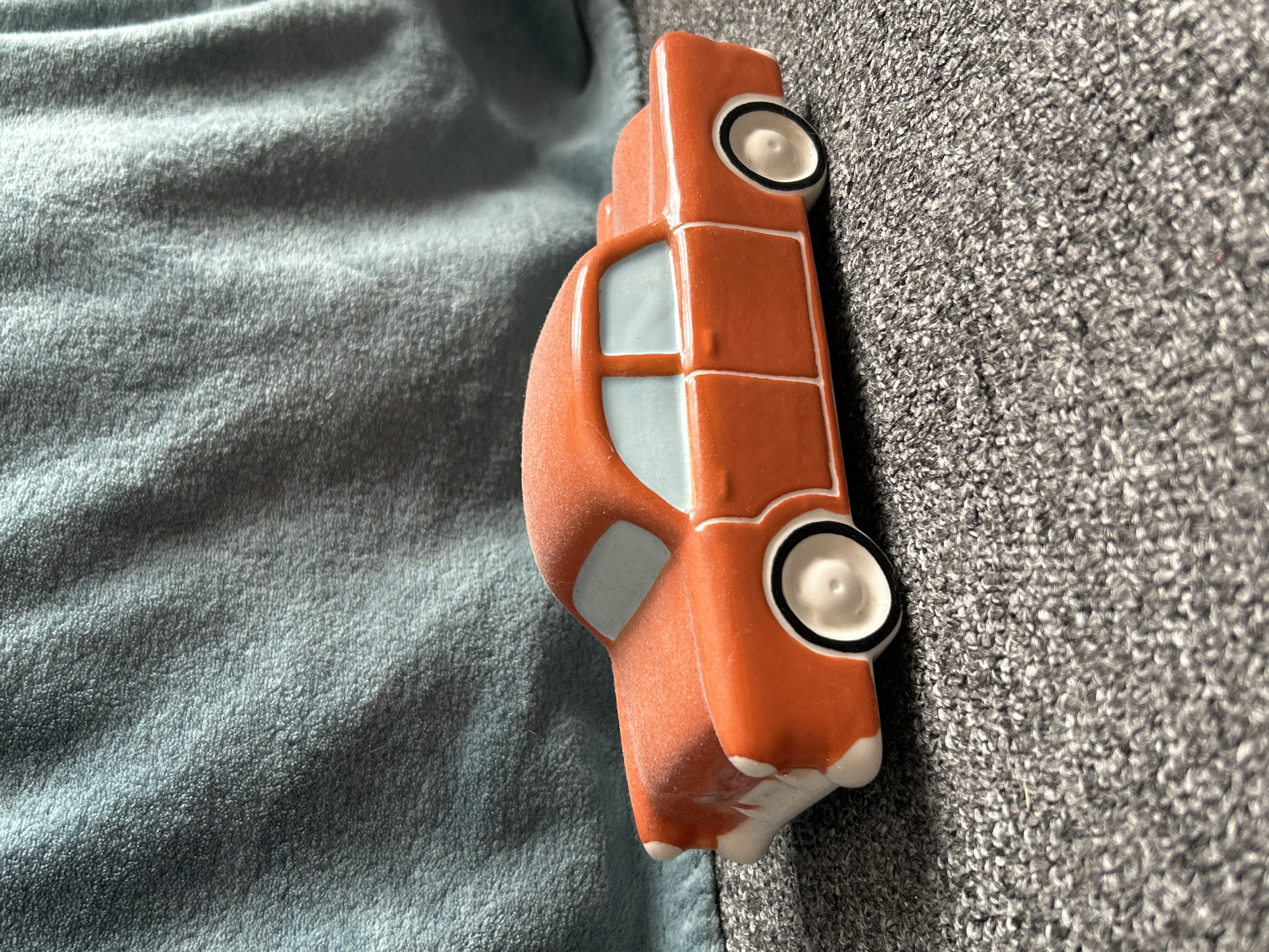 An orange ceramic toy car with white trim and light blue windows sits on a gray textured surface. Behind the car, a crumpled teal-colored fabric creates a backdrop. The scene is lit from the right, casting a slight shadow to the left of the car. There are no people, actions, or interactions depicted. The image does not provide any information about its location in Moscow, Russia.