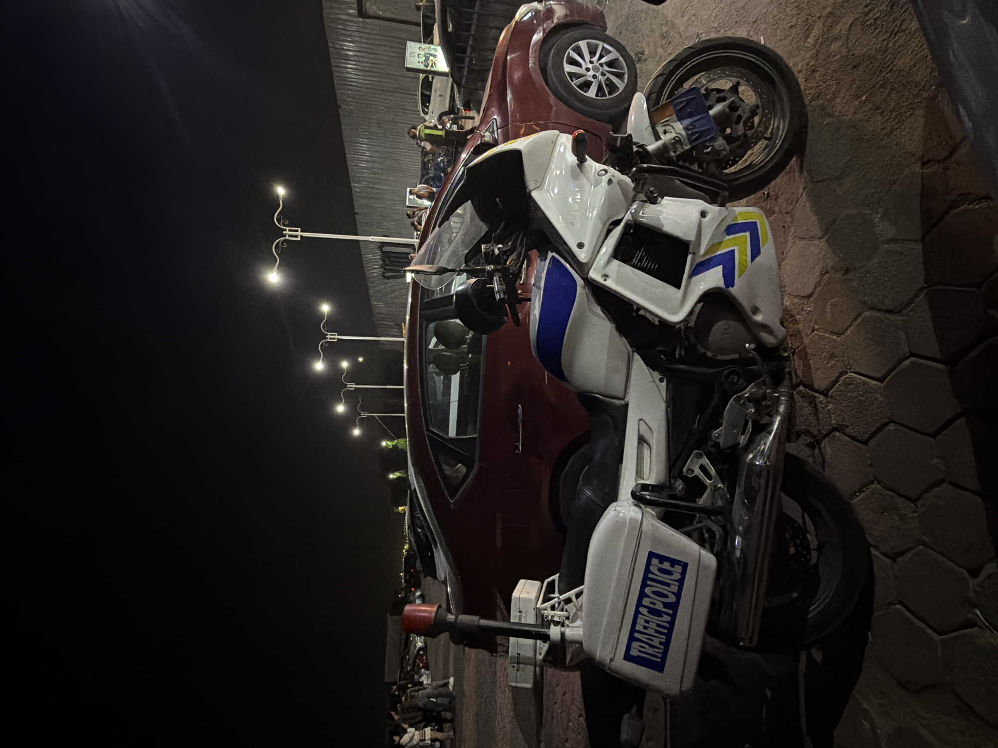A traffic police motorcycle is parked at night on a textured paved surface in Sharm El Sheikh, Egypt. The motorcycle is white with blue and yellow markings and has "TRAFFIC POLICE" written in blue on its side cases. A red flashing light is mounted on top of the motorcycle. A red sedan is parked behind the motorcycle, and other cars are visible in the background. Several people are gathered near the cars, and streetlights illuminate the scene. The background also shows a corrugated metal fence.