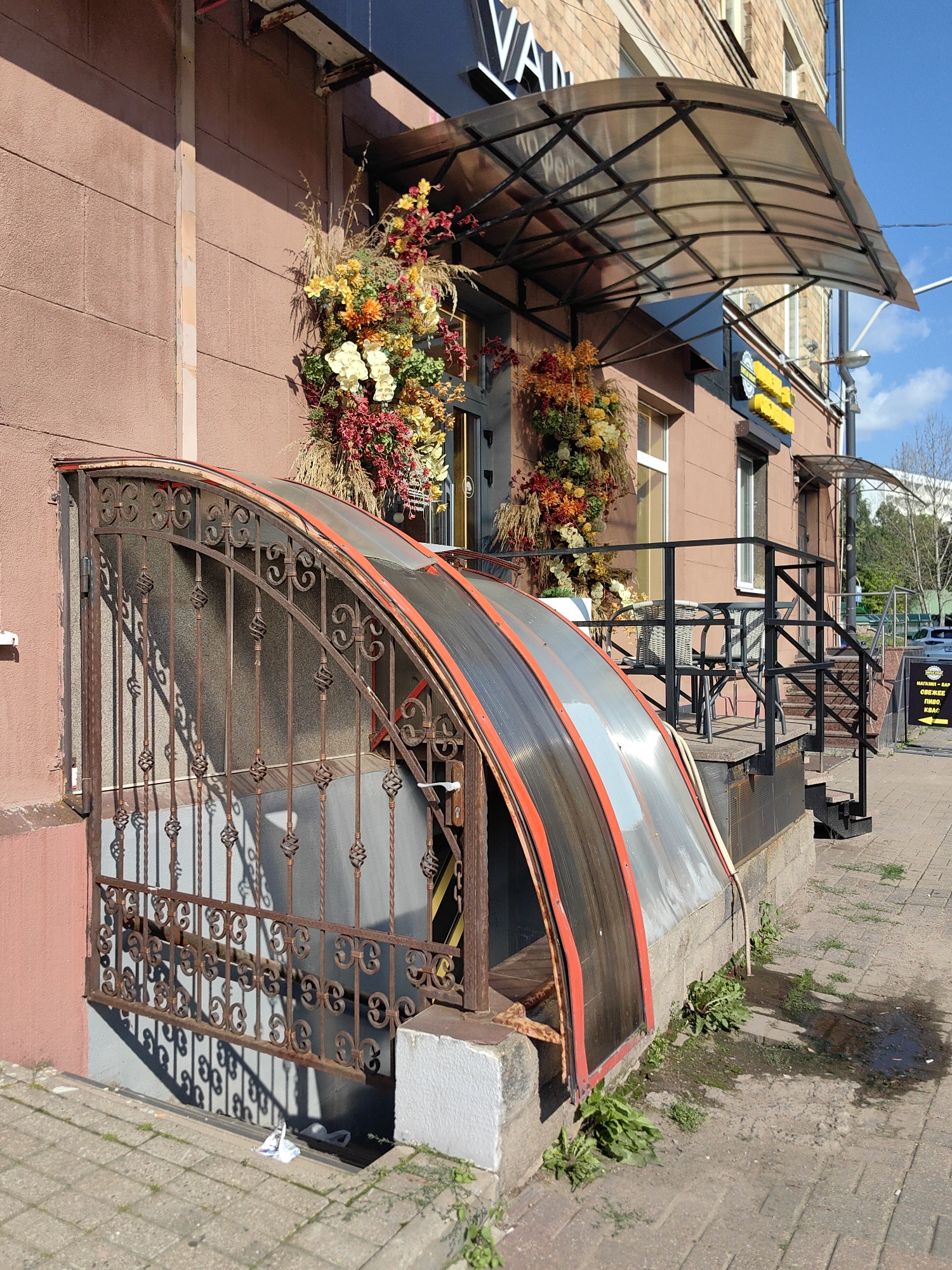 The image shows the entrance to a building in Minsk, Belarus, likely a cafe or shop, with a visible sign that says "VAN...". The entrance features a decorative metal gate covering a basement-level entryway. Above this is a modern, arched plastic and metal awning. The building itself has a reddish-brown textured facade and is a multi-story structure with brickwork visible on the upper floors. Large floral arrangements, featuring autumnal colors like yellow, orange, and red, are prominently displayed near the entrance. To the right, there are steps leading up to an elevated area with outdoor seating, including tables and chairs. The ground is paved with brickwork, and there is evidence of vegetation growing around the building's foundation. The scene appears to be a sunny day with shadows cast on the ground.