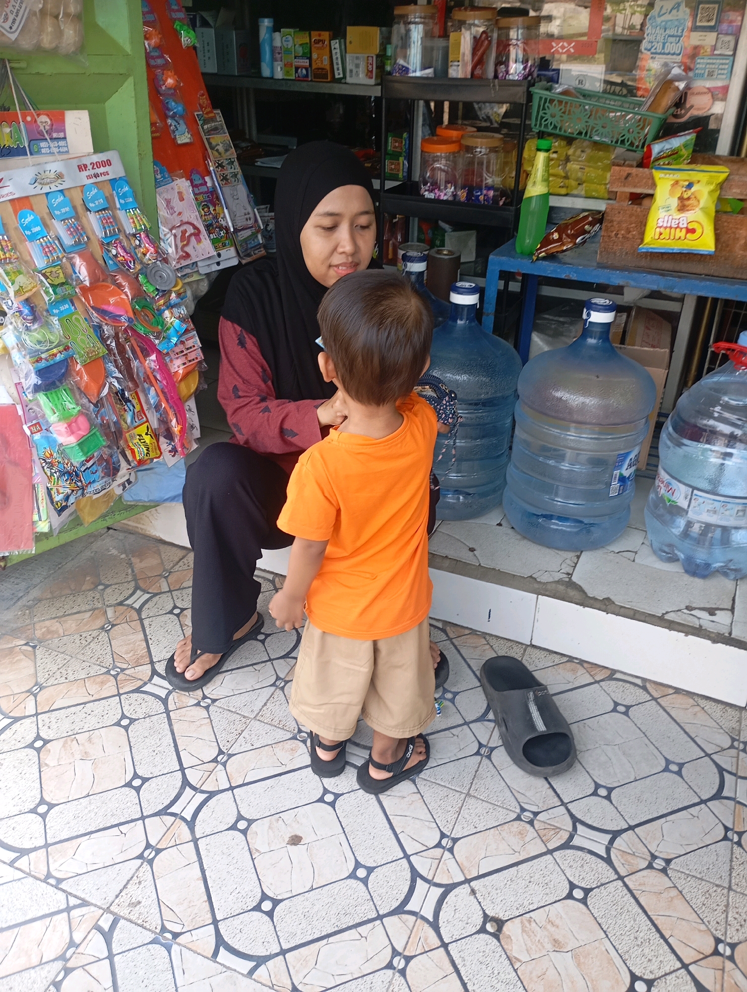 A woman wearing a black hijab and a maroon and black patterned top is sitting on the ground, facing away from the camera. She is looking down at a young boy who is standing in front of her, also facing away from the camera. The boy is wearing an orange t-shirt and light brown shorts. The scene appears to be in front of a small shop or stall, possibly in Babelan, Indonesia. To the left of the woman, there are shelves displaying various items, including packets of snacks and toys. In front of the shop, there are several large, clear water jugs. The ground is tiled with a patterned design. To the right of the boy, there is a black sandal lying on the ground.