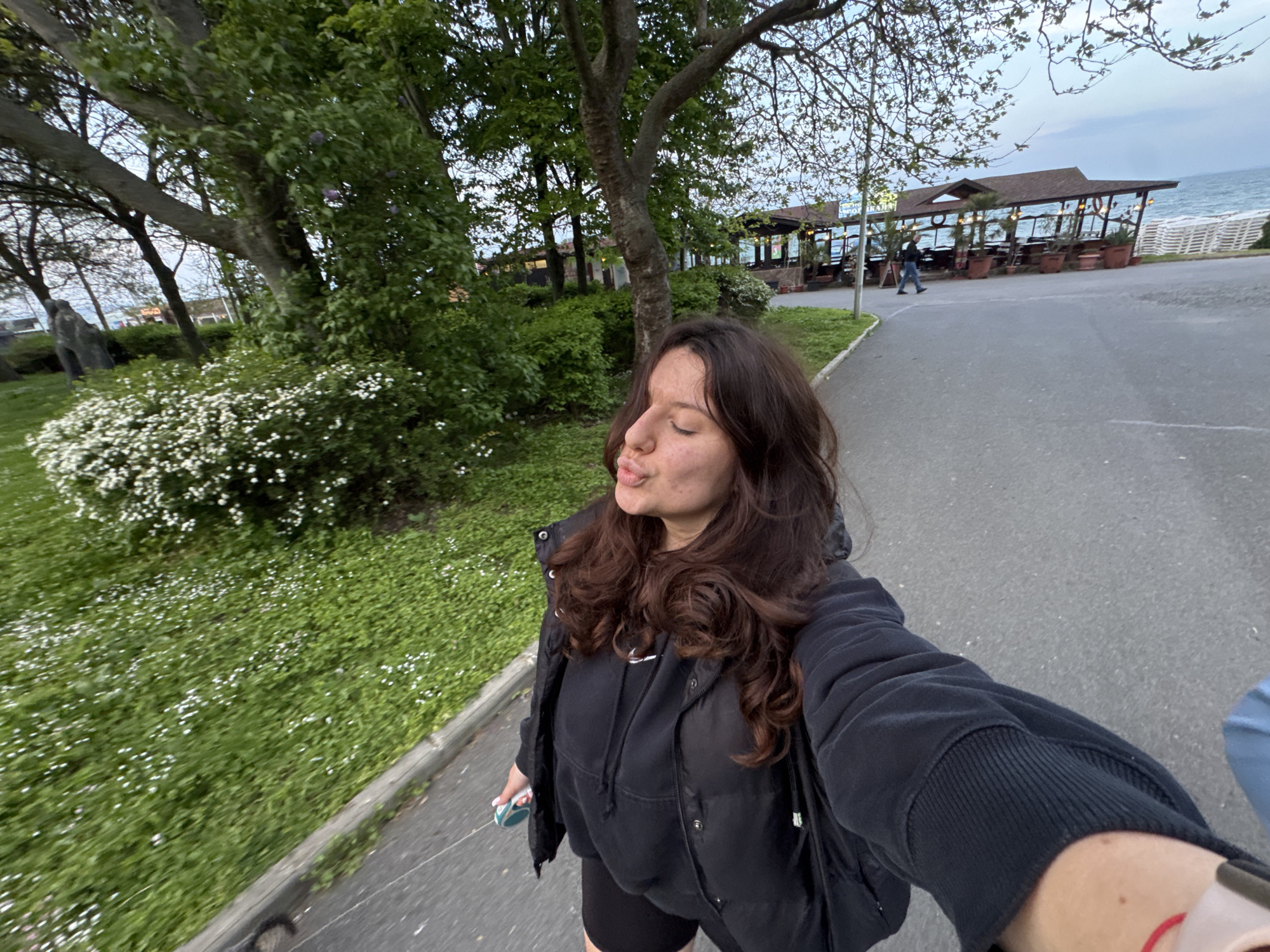 A young woman takes a selfie on a paved path, likely in Burgas, Bulgaria, given the presence of a beachfront restaurant structure in the background reminiscent of the city's seaside establishments. She is wearing a black hooded sweatshirt, a puffy vest, and black shorts, and she is holding a small item in her left hand. The woman purses her lips and closes her eyes. To her left is a grassy area with small white flowers and a line of trees. The restaurant structure is visible in the distance and includes a roof and open sides with tables and chairs visible inside. To the right of the structure is the sea. Other people are walking in the background.