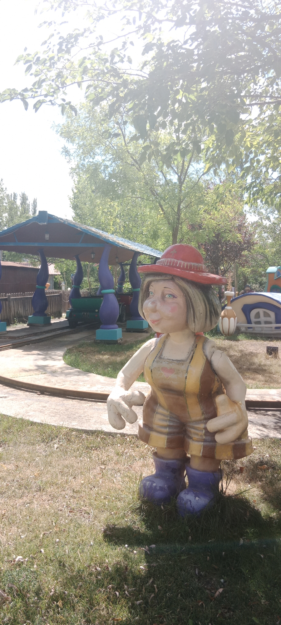 In Torrejón de Ardoz, Spain, this image captures a section of an amusement park. The foreground features a life-sized, cartoonish statue of a woman with a wide smile, wearing a red hat and a striped yellow and brown jumpsuit with blue boots. Her large hands are positioned as if gesturing. Behind the statue, a whimsical amusement park ride is visible. This ride consists of a blue and purple canopy supported by intricately designed pillars. A small green vehicle, likely part of the ride, is situated on a curved track beneath the canopy. The background reveals more of the park's attractions, including colorful structures resembling small houses, and lush green trees overhead. The ground is covered in a mix of dry grass and dirt, indicating a sunny outdoor environment.