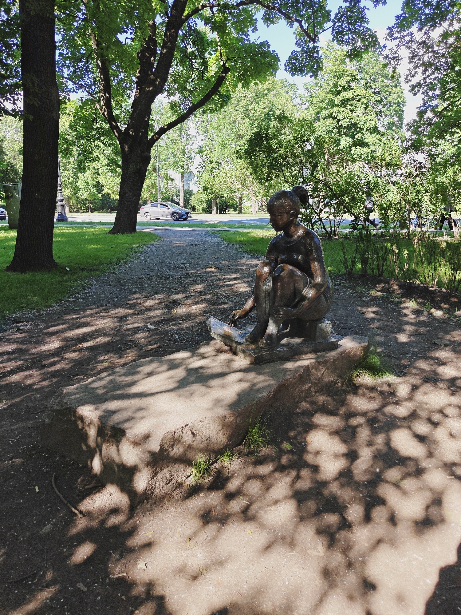 A bronze statue of a young girl in a seated position is the central subject of the image, located in a park-like setting. She is positioned on a rough-hewn stone base, with her legs drawn up and her arms resting on her lap. She appears to be interacting with a flat object in front of her, possibly a book or a slate. The statue is casting a shadow onto the stone base and the surrounding dirt path.
The environment suggests a sunny day with dappled sunlight filtering through the leaves of mature trees that surround the area. A gravel path leads away from the statue, curving to the left and disappearing into the background. Beyond the path, a well-maintained grassy area is visible, with a street and cars in the distance. Lush green trees and bushes form a dense backdrop, creating a peaceful and natural atmosphere. The presence of a street and trees indicates an urban park setting.
While not explicitly stated, the style of the statue, the urban park landscape, and the architectural elements visible in the background (streetlights, potentially buildings obscured by foliage) are characteristic of Saint Petersburg, Russia. The statue likely depicts a scene from childhood or a literary figure, common themes in public art found in the city's numerous parks and gardens.