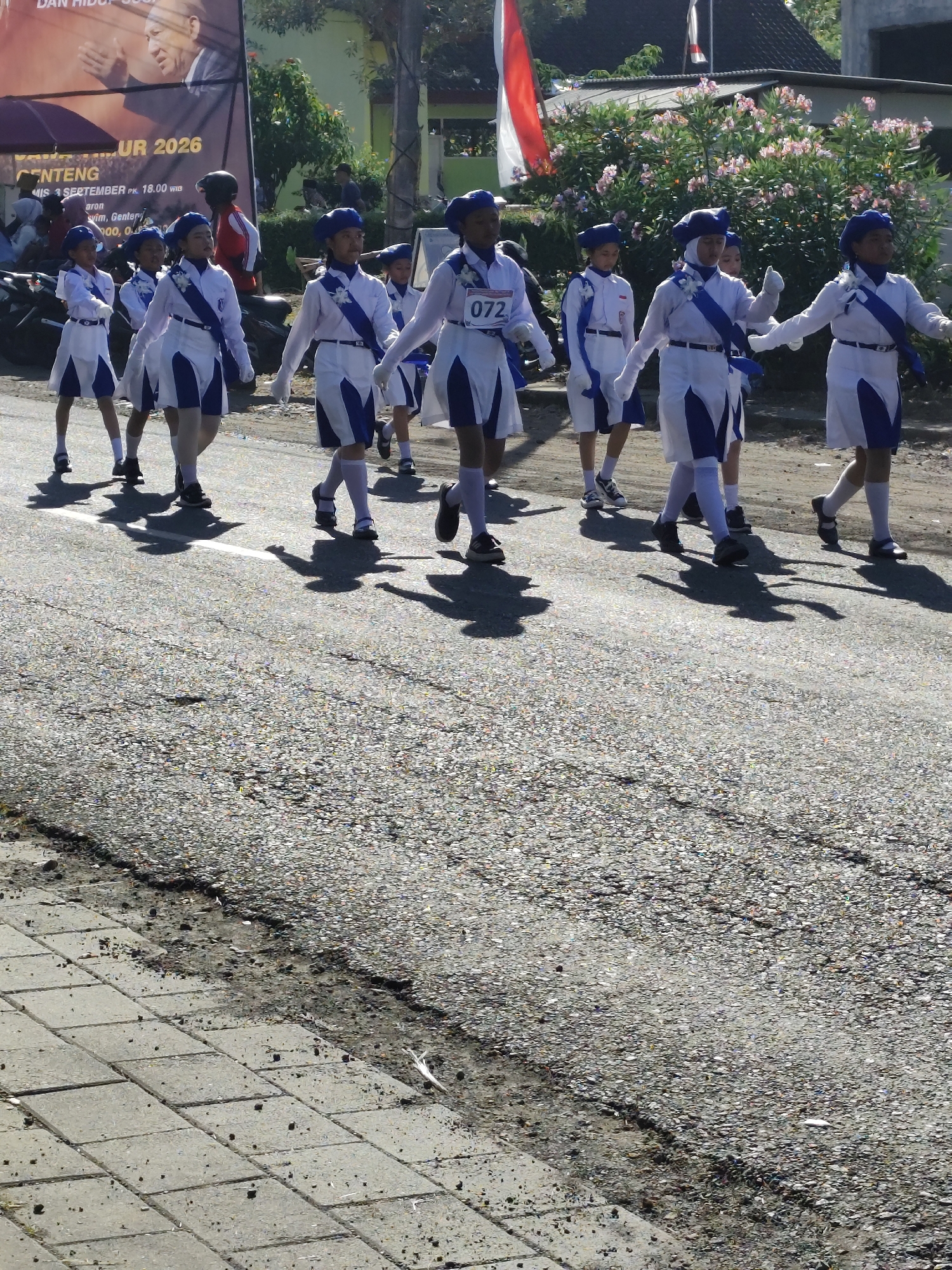A group of girls in matching blue and white marching band uniforms walk in formation along a cracked asphalt road. The girls wear blue hats, white long-sleeved shirts with blue accents, white skirts, white gloves, and white knee-high socks. Some have blue sashes across their chests. They are walking from left to right in the frame, casting long shadows on the road. In the background, there is a large banner with text that includes "UR 2026" and "GENTENG," and a smaller banner with a red and white Indonesian flag. There are also buildings and trees in the background. The scene appears to be outdoors during the daytime. The specific location is the city of Tegalarum, Indonesia, as indicated by the banner text.