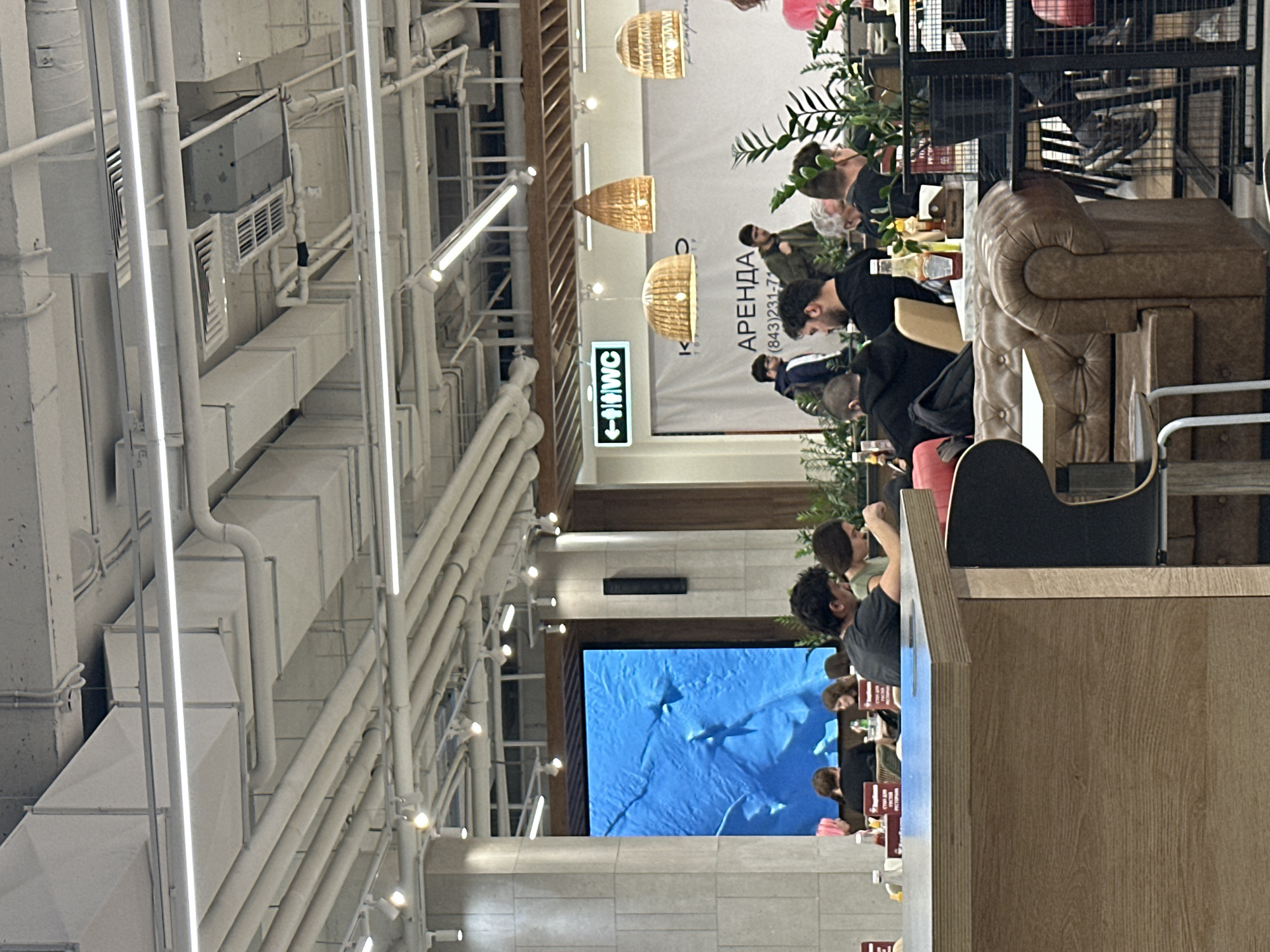 The image depicts an interior setting, likely a shopping mall or food court in Kazan, Russia, as indicated by the text "АРЕНДА КАЗАНЬ" and a phone number with the country code +7 (843). Several people are seated at tables, some appearing to be engaged in conversation. A large screen displays an image, possibly of sharks in an ocean. Overhead, the ceiling is exposed, revealing a complex network of pipes and ventilation systems. Wicker pendant lamps hang from the ceiling above the seating area. A WC sign is visible.