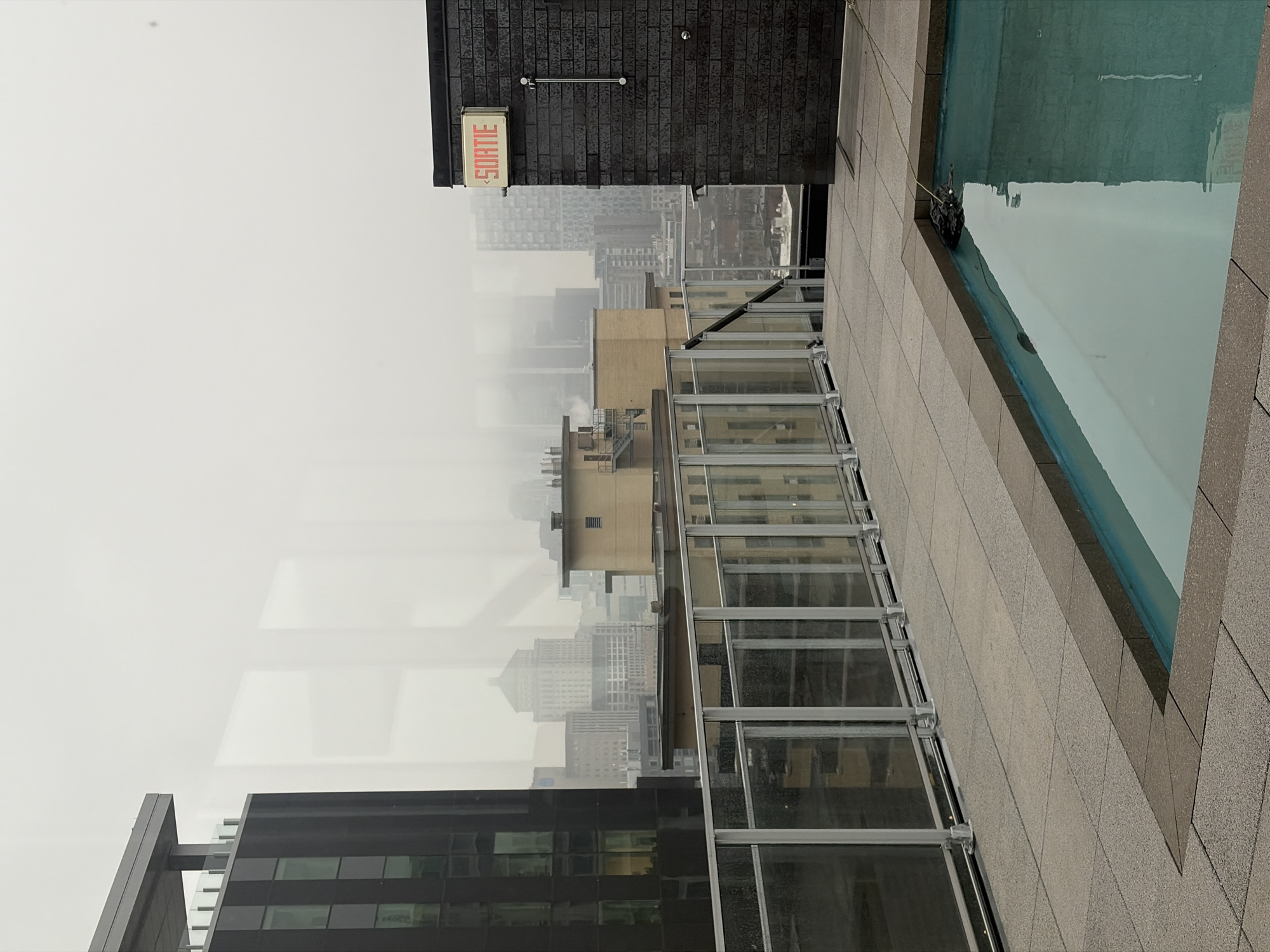 The image displays a rooftop scene overlooking a city, likely Montreal, Canada, under very foggy or heavily overcast conditions. In the foreground, occupying the right side, is a rectangular swimming pool filled with light blue water, surrounded by light-colored paving stones. A dark object, possibly a pool cleaner or a cover weight, is visible in the water near the edge, attached to a light-colored rope or cord.
A modern glass and metal railing runs across the middle of the frame, separating the pool area from the city skyline. Beyond the railing, the urban landscape unfolds, but most buildings are heavily obscured by the dense fog or clouds, making their details indistinct. Closer to the foreground on the left, part of a building with dark glass windows is visible.
Above the city view, towards the upper left, a dark brick wall is prominent. Mounted on this wall is a cream-colored rectangular sign bearing the French word "SORTIE" (meaning "EXIT") in red capital letters. Below the sign, a horizontal metal bar and two small round fixtures are attached to the brickwork. The overall impression is one of a quiet, hazy, and cool daytime atmosphere. No individuals or specific activities are depicted in the static scene.