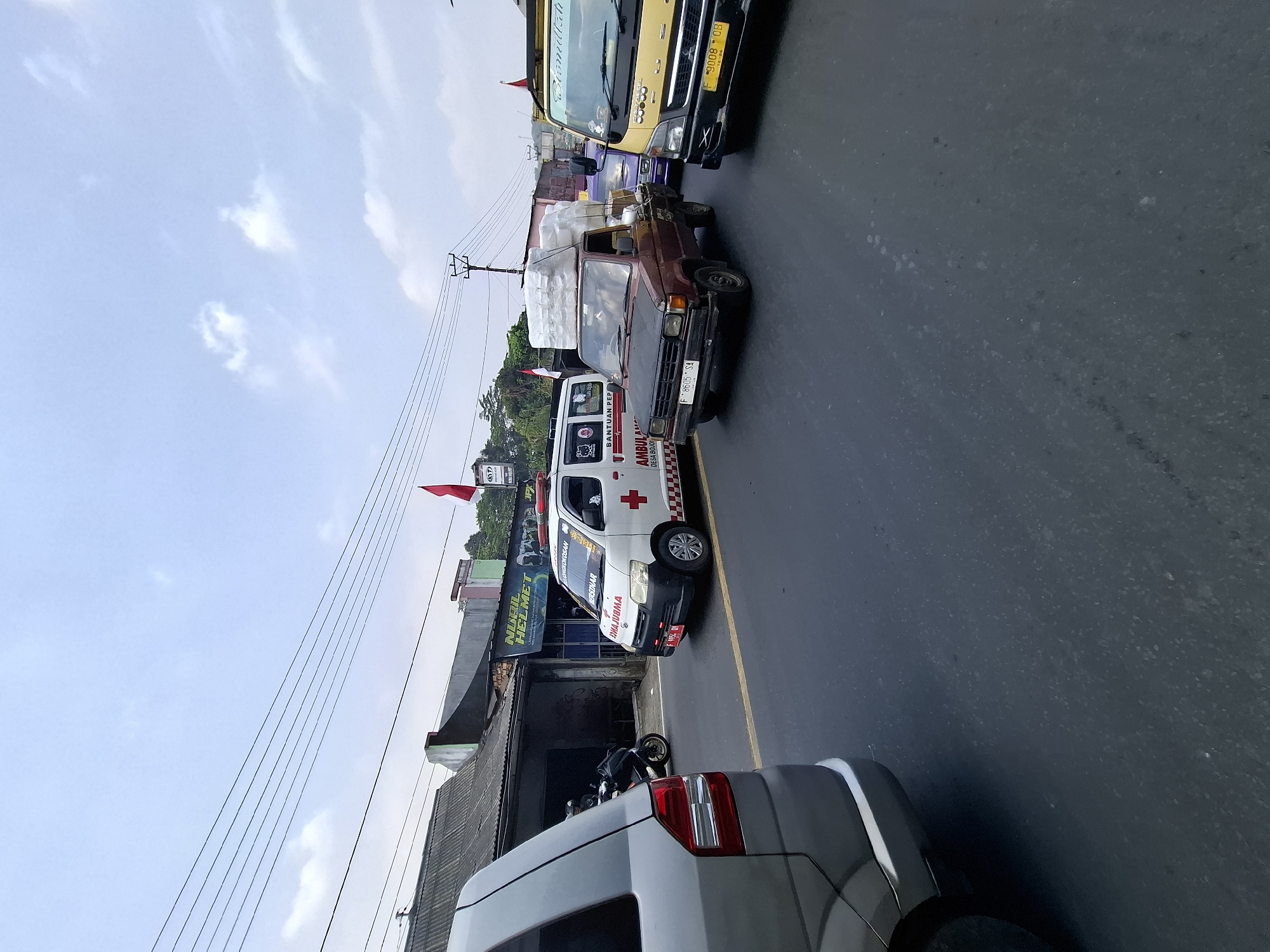 The image depicts a traffic scene on a road in Pamuruyan, Indonesia. A grey minivan is parked on the left, facing away from the camera. In the middle of the road, an ambulance is driving, and to its right, a brown pickup truck with a load of white goods is also moving. Further to the right, a yellow truck is visible, and behind it, a purple car. The road is paved and marked with a yellow line. Several buildings are situated along the side of the road, with some signs visible, including one for "NURIL HELMET". The sky is partly cloudy. Power lines are strung overhead, supported by utility poles. A red and white Indonesian flag is attached to a pole near one of the buildings. A motorcycle is parked in front of one of the buildings.