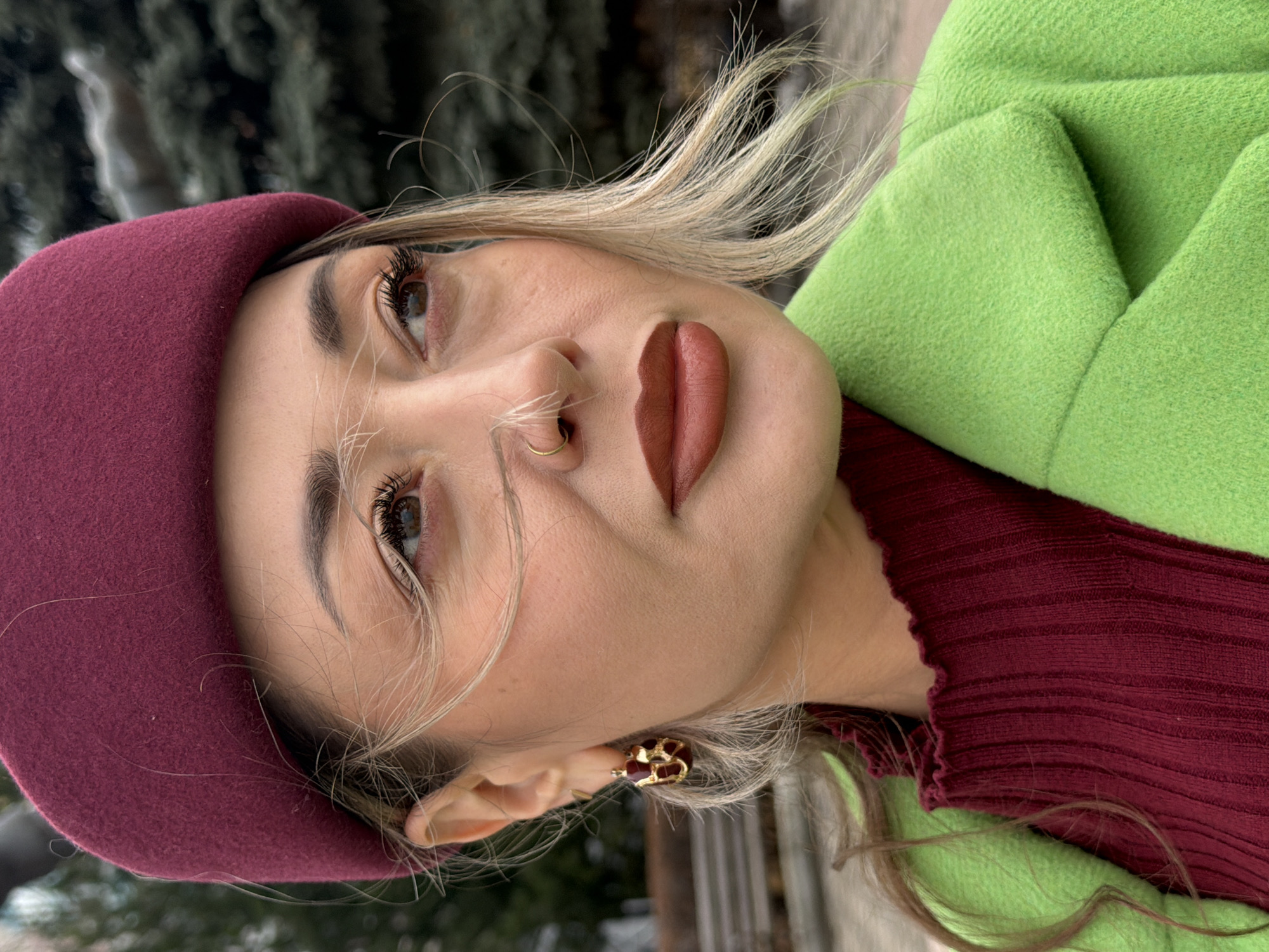 A woman is facing the camera at a slightly tilted angle. She is wearing a burgundy beret, a burgundy ribbed sweater, and a light green coat. She has blonde hair and is wearing a gold nose ring and gold earrings. The background appears to be outdoors, possibly a park, with trees visible. Due to the lack of identifiable landmarks, it is impossible to confirm the specific location within the city of Krasnoyarsk, Russia.