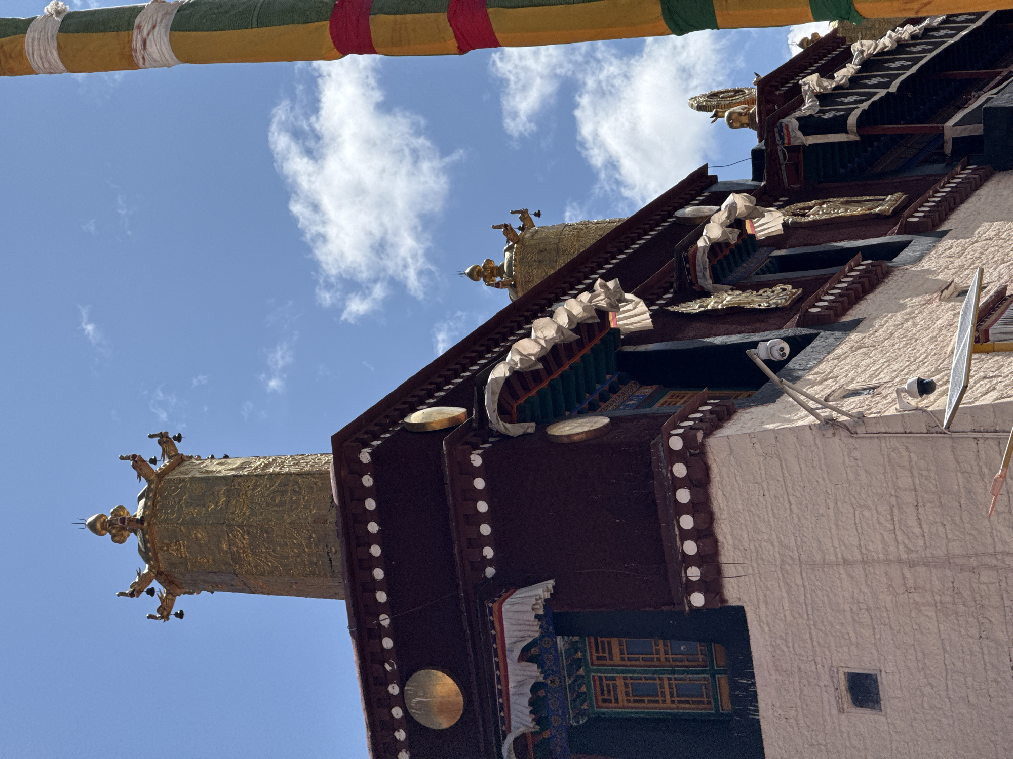 The image shows a building in Lhasa, China, with traditional Tibetan architectural elements. The building is multi-tiered with a white facade and dark red and gold accents. A large, golden cylindrical structure with decorative elements is attached to the building. Above the building, the sky is visible with scattered clouds. At the top of the image, a colorful striped cloth is draped across the scene. A security camera is mounted on the building's facade.