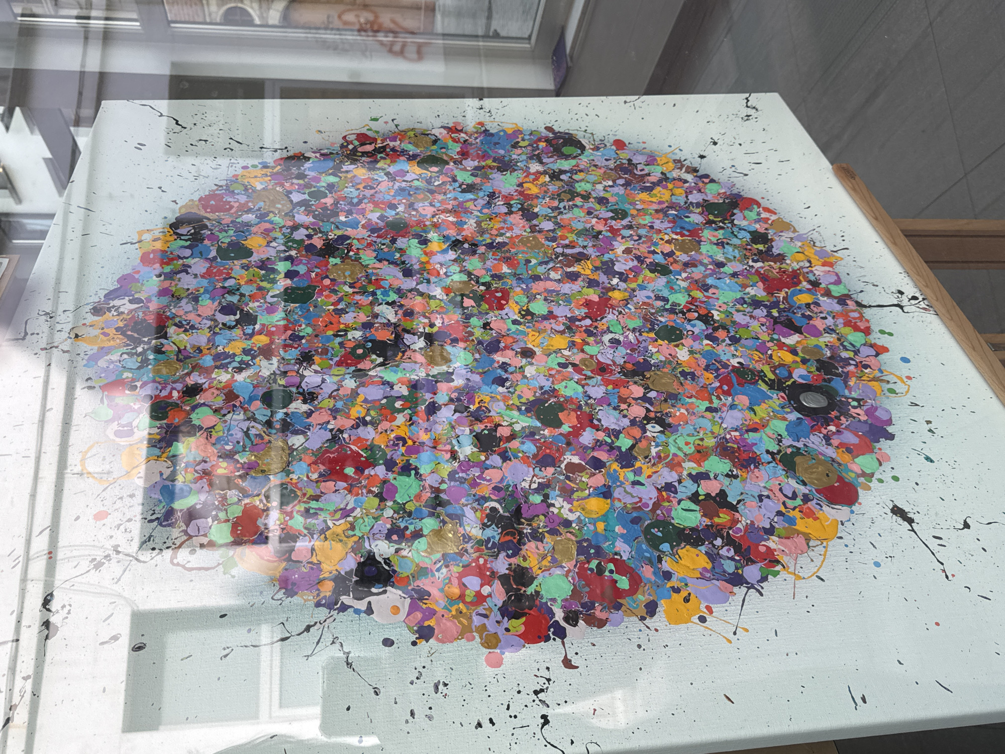 A close-up, angled shot captures a brightly colored, abstract painting on a canvas displayed on a wooden easel. The painting features a dense concentration of small, circular splatters of various vibrant hues, including red, blue, green, yellow, purple, and pink, with some gold accents. The splatters are layered and create a circular, mandala-like pattern that dominates the canvas. Black paint splatters are also visible, extending outwards from the central design onto the pale blue background of the canvas and also on the glass fronting the painting. The artwork is presented in what appears to be a shop window, with reflections of buildings and the street visible on the glass. The setting suggests an urban environment, possibly Antwerp, Belgium, given the architectural style suggested by the reflections. The image focuses on the texture and vibrant chaos of the artwork, with minimal background detail.