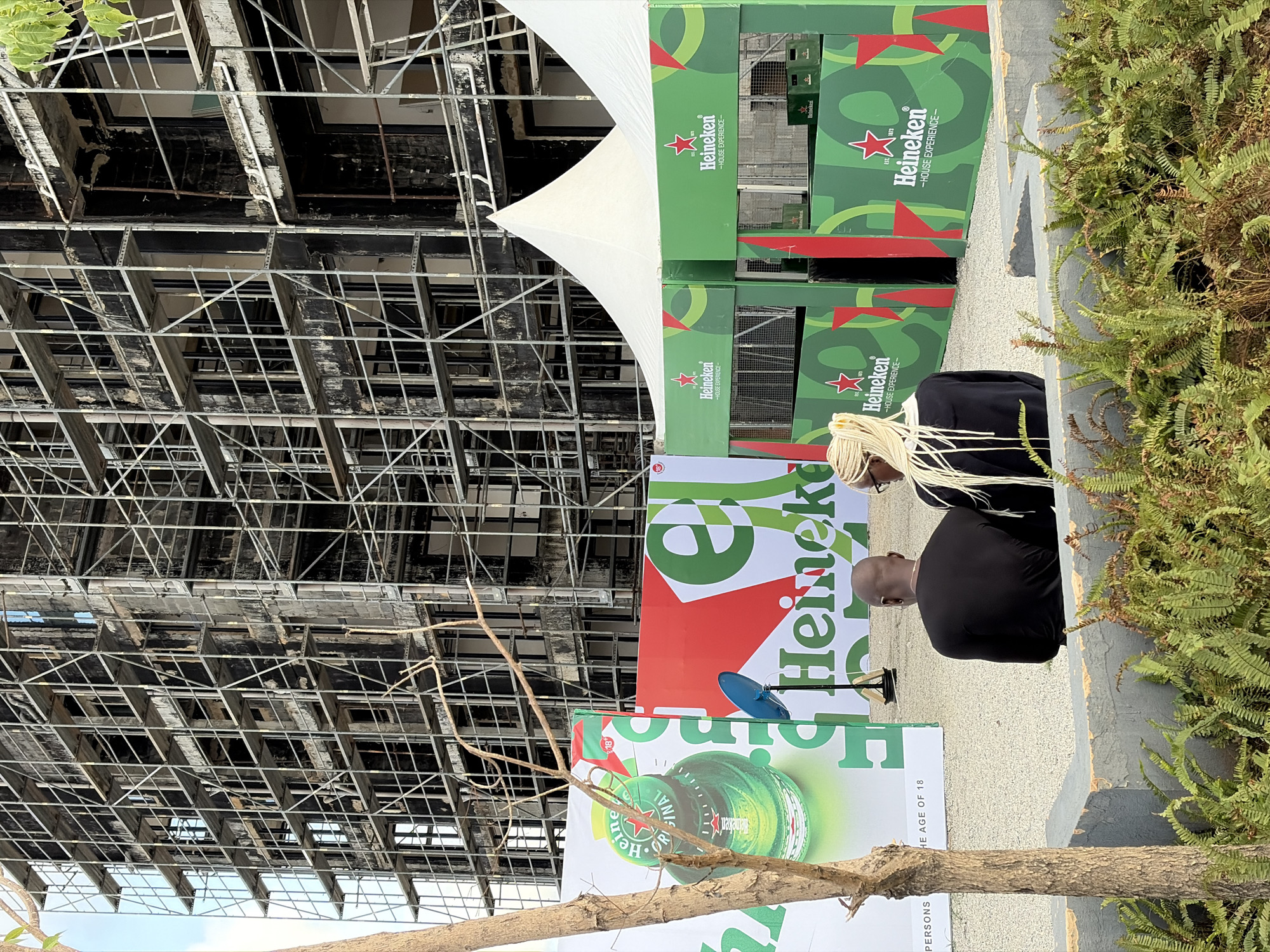 The image depicts an outdoor scene in Lagos, Nigeria, dominated by a partially destroyed building and a Heineken promotional event. The building behind the event is characterized by a skeletal framework, indicating severe damage or demolition. In the foreground, there's a Heineken-branded setup, including posters, tents, and green-themed decor, which suggests a marketing or event space. Two individuals are visible within the event area. Ferns and foliage on the right suggest a somewhat landscaped environment.