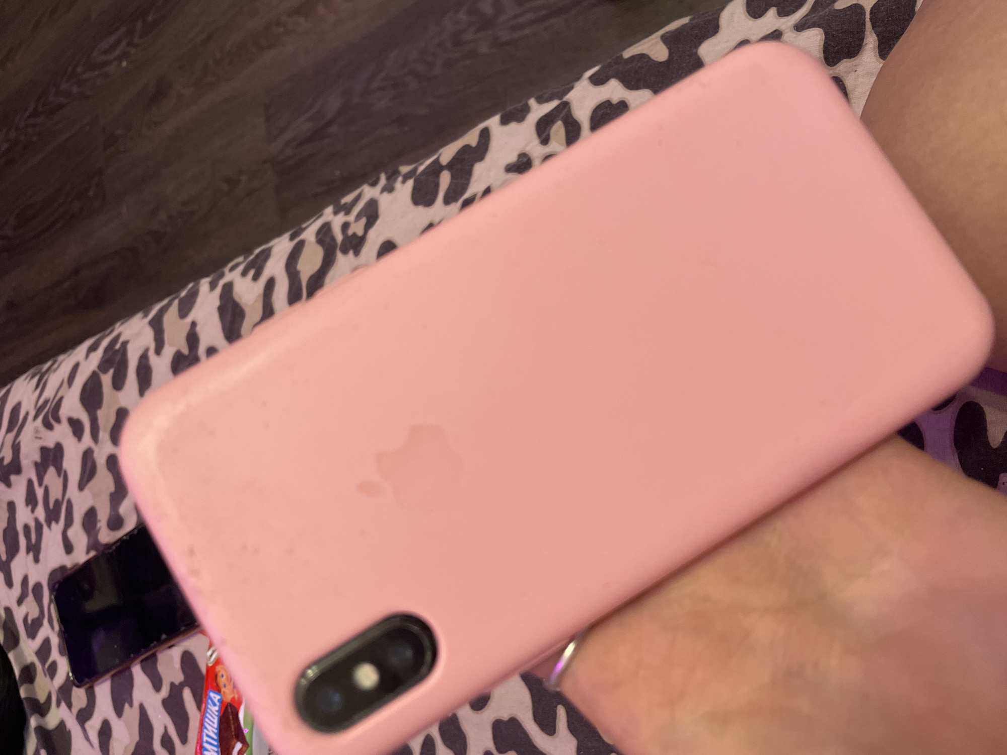 In Ladozhskoe Ozero, Russia, a pink iPhone case with a visible Apple logo lies on a patterned surface. To the left of the iPhone case, a partially obscured black smartphone is visible, as well as a candy wrapper with Russian text. The patterned surface appears to be fabric with a cow print design. The overall scene is indoors, likely a domestic setting.