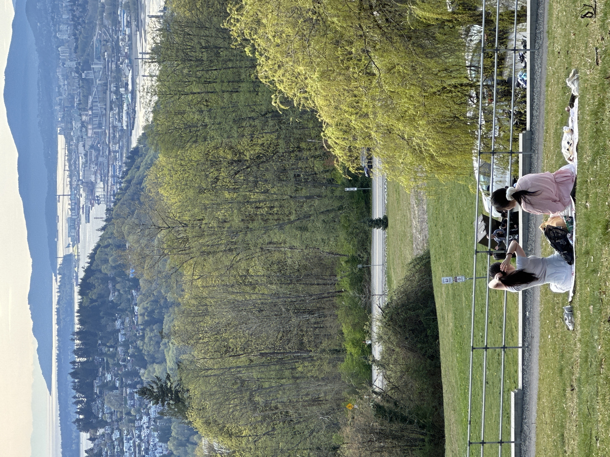 The image depicts a scenic view in the District of North Vancouver, Canada. From an elevated vantage point, two individuals are visible relaxing on a blanket laid on the grass, near a metal railing. One person is wearing a pink dress, while the other is dressed in a light-colored t-shirt and shorts. The landscape features lush greenery with a dense forest that extends toward the city and coastline in the distance. The cityscape consists of buildings and industrial structures, with water bodies separating areas of land, against a backdrop of distant mountains. A road cuts through the green spaces near the foreground.