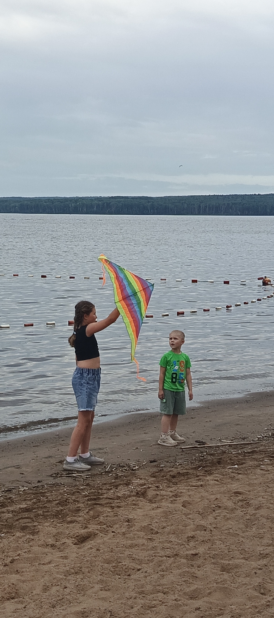 Two children are on a sandy beach, with a large body of water behind them. The water has a rope with buoys marking a swimming area. A dense forest lines the opposite shore of the water. The sky is overcast with clouds.
In the foreground, on the left, a young girl with braided brown hair stands facing right, holding a colorful rainbow kite above her head. She is wearing a black crop top and denim shorts. On her feet are grey sneakers.
To the right of the girl stands a young boy, facing left. He is wearing a green t-shirt with a graphic design and white shorts. He has on white socks and light-colored sneakers.
The scene is located on the beach of the Kama River in Chaikovsky, Russia.
