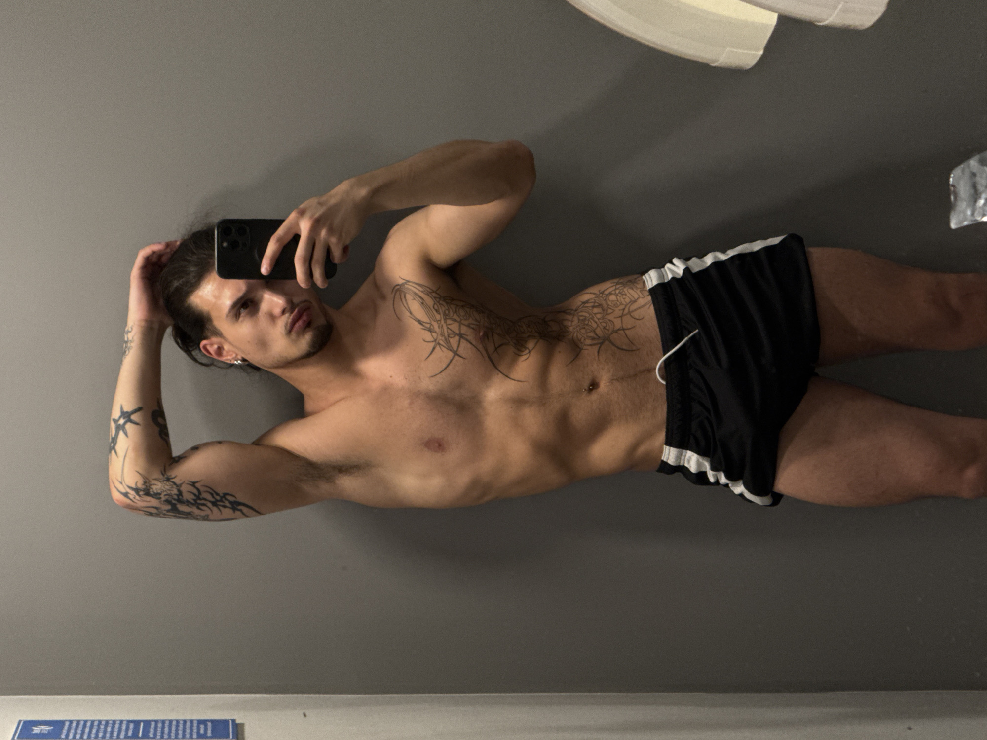 In a dimly lit changing room with plain grey walls, a shirtless young man with dark hair pulled back in a bun takes a mirror selfie. He is wearing black athletic shorts with white stripes on the sides. He has tattoos on his left arm and torso. His right arm is raised, touching his head, and his left hand holds a black smartphone. A white sign with blue text is visible on the left wall.