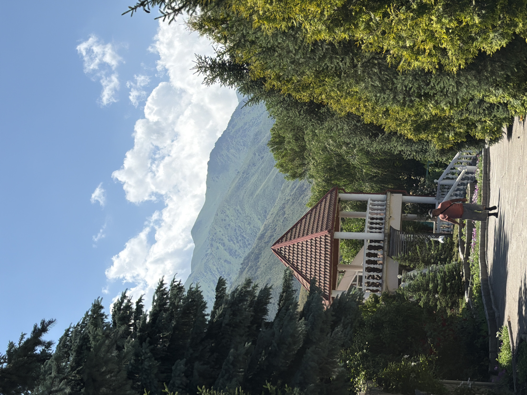 A man walks on a paved path in a park on a sunny day. He is wearing an orange t-shirt and grey shorts. To his left, a gazebo with a red tiled roof and white balustrades is visible. The park is surrounded by lush green trees and in the background, a mountain range is visible under a bright blue sky with white clouds.