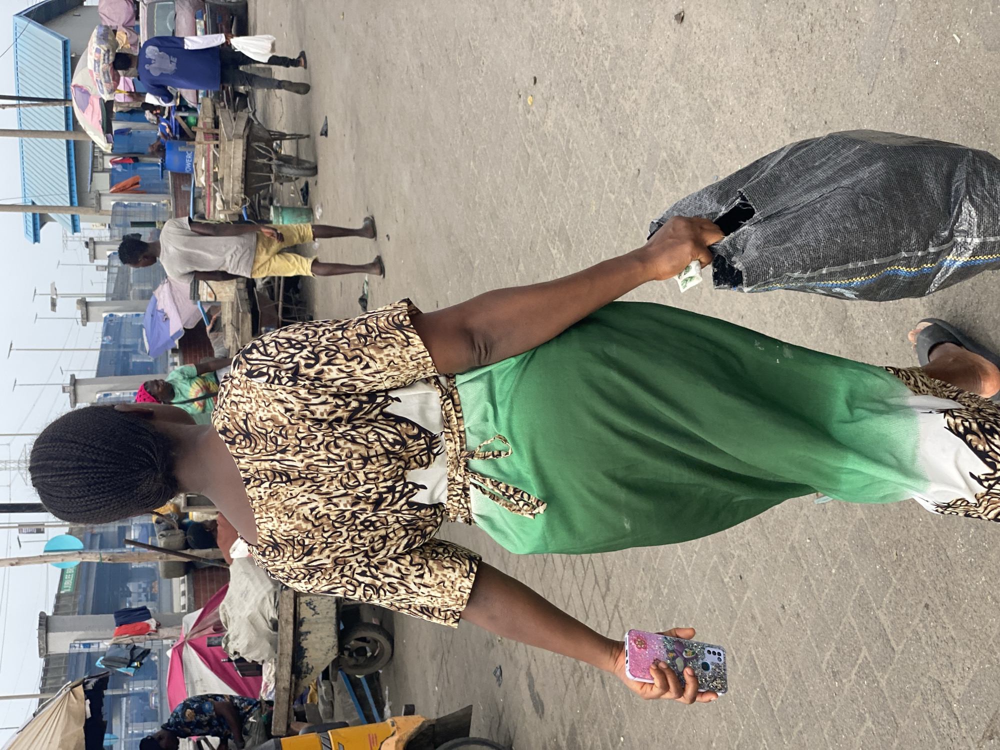 The image shows a woman in Lekki, Nigeria, standing on a concrete surface typical of a public area. She is wearing a patterned top, a green skirt, and sandals. She holds a black bag in one hand and a smartphone in the other. The background reveals a busy street scene with stalls, people, umbrellas, and parked vehicles.