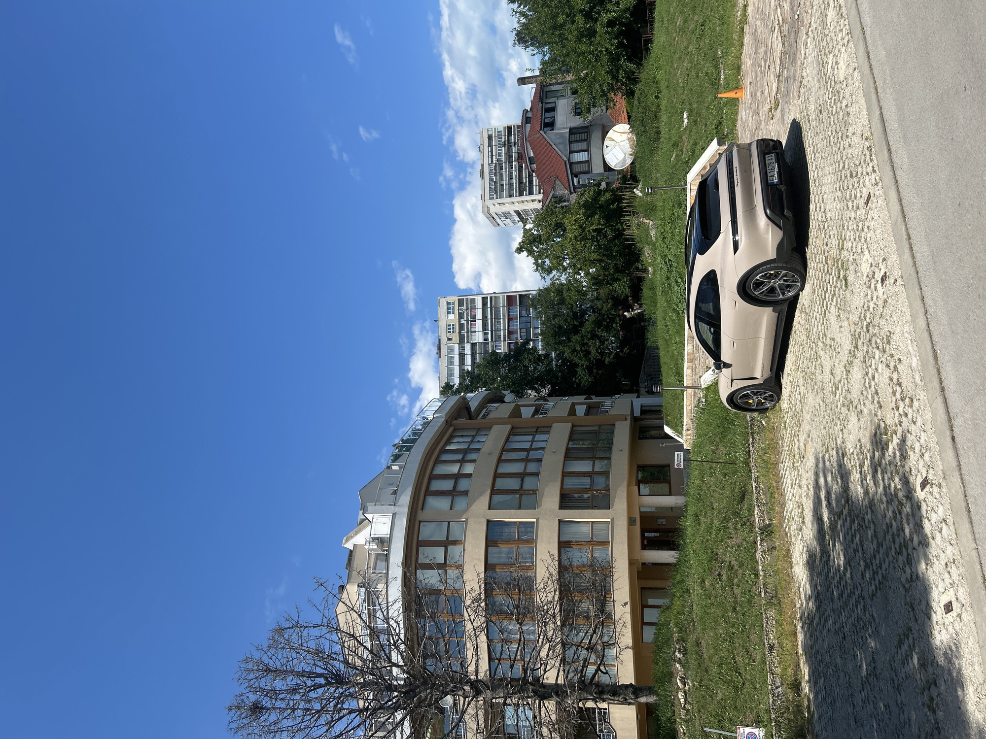 The image shows a street scene in Varna, Bulgaria, on a sunny day with a clear blue sky and scattered white clouds. In the foreground, a beige car is parked on a cobblestone surface next to a patch of grass. The car has tinted windows and a license plate with the text "EM678AA". Behind the car, a building with a curved facade and large windows is visible, along with a bare tree in front of it. In the background, several other buildings, including high-rise apartments, are seen among lush green trees. A satellite dish is mounted on one of the buildings. The overall impression is a typical urban environment on a pleasant day. No people are visible in the image. There are two signs near the car, but the text on them is not legible. The sun is casting shadows, suggesting it is daytime, likely late morning or early afternoon.