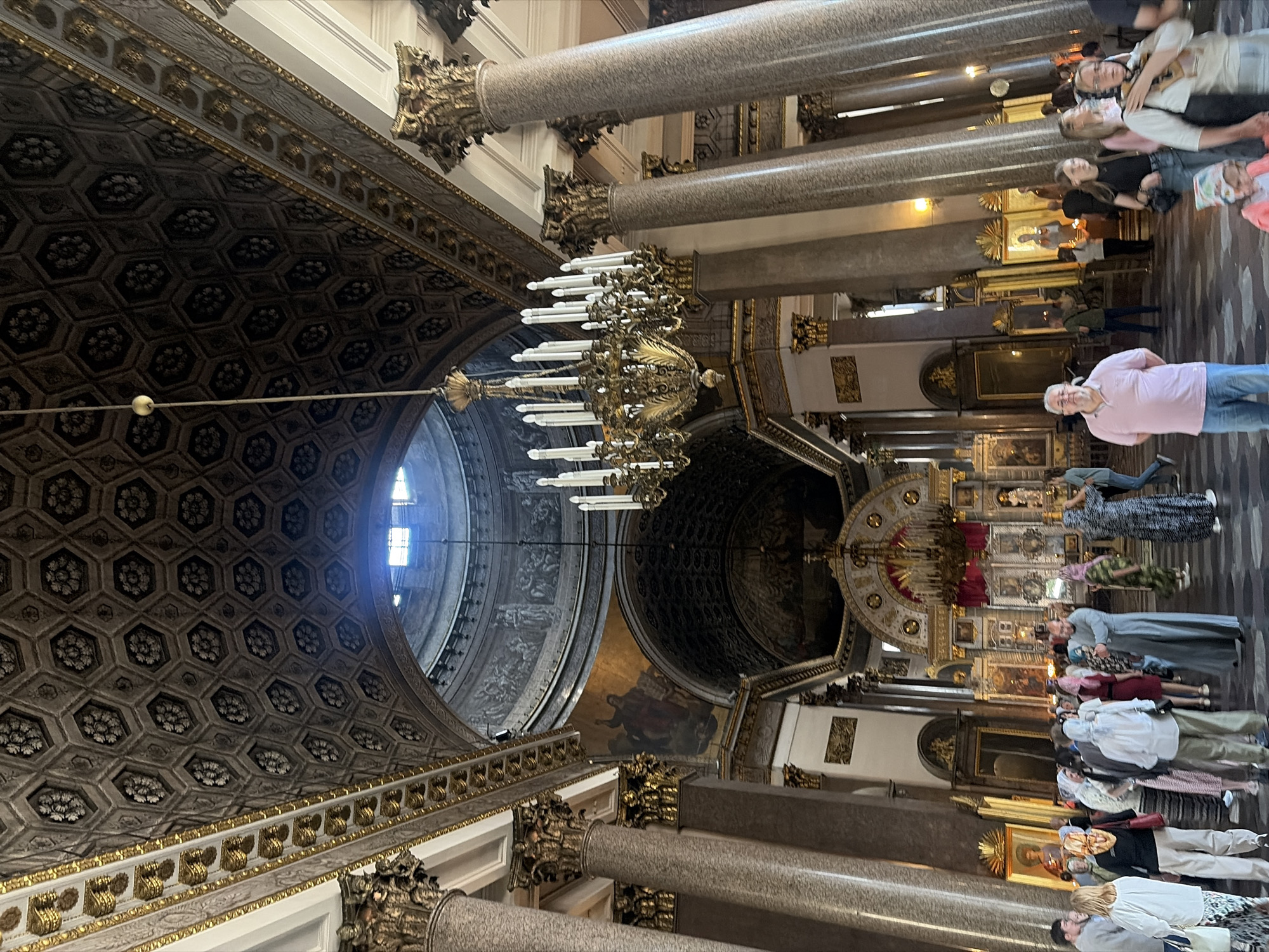 The interior of a grand cathedral is depicted, characterized by a high, intricately decorated ceiling with hexagonal patterns and floral motifs. Ornate gold trim runs along the edges of the ceiling and adorns the tops of massive, fluted columns that support the structure. A large, multi-tiered chandelier hangs prominently in the center, featuring numerous candle-like fixtures. Natural light streams in from a large opening in the dome, illuminating the scene and highlighting the architectural details. Religious artwork, including mosaics and icons, is visible on the walls and behind an altar area in the background, which is adorned with red drapery and further iconography. A group of people, likely tourists and worshippers, are gathered throughout the space, observing the architecture and religious artifacts. Some are dressed in traditional headscarves, common for visitors to religious sites. The floor is a dark, reflective material. The overall impression is one of opulence and religious significance.